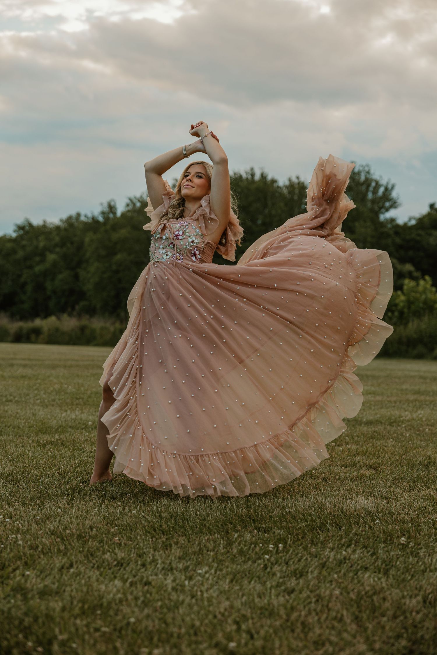 Woman twirling in a flowing pink tulle ballgown on a green lawn with trees and cloudy sky.