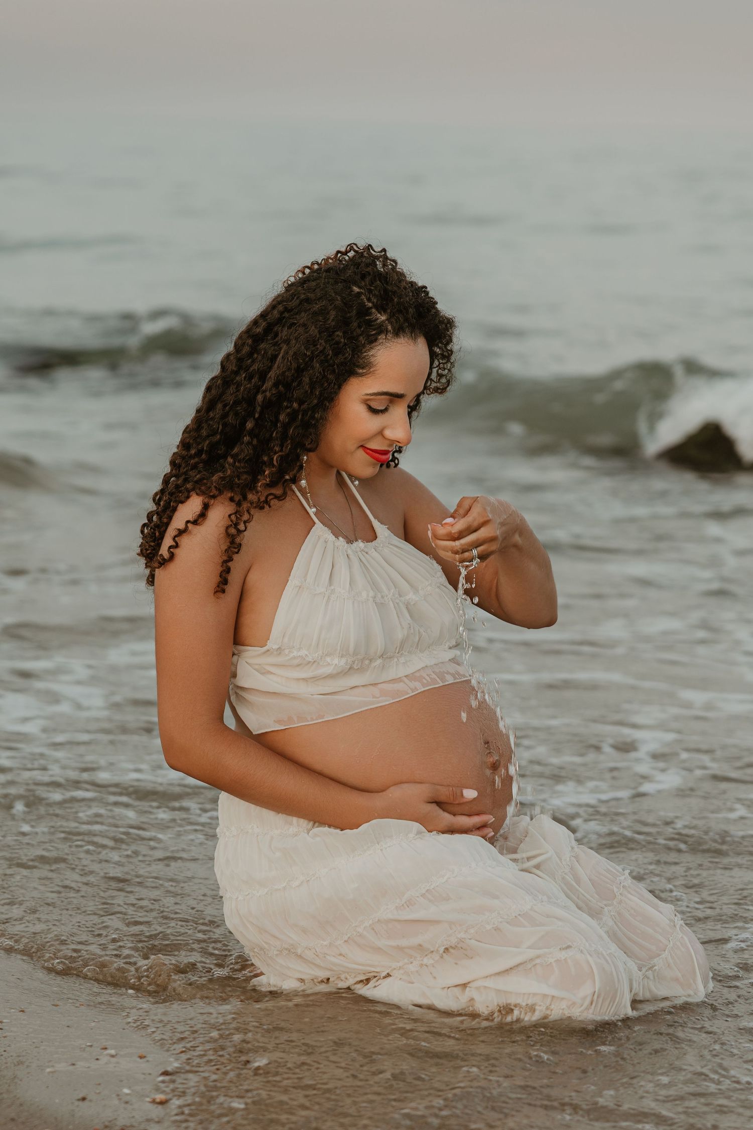 Pregnant woman in white dress sitting at ocean's edge, cradling her baby bump on a serene beach at dusk.