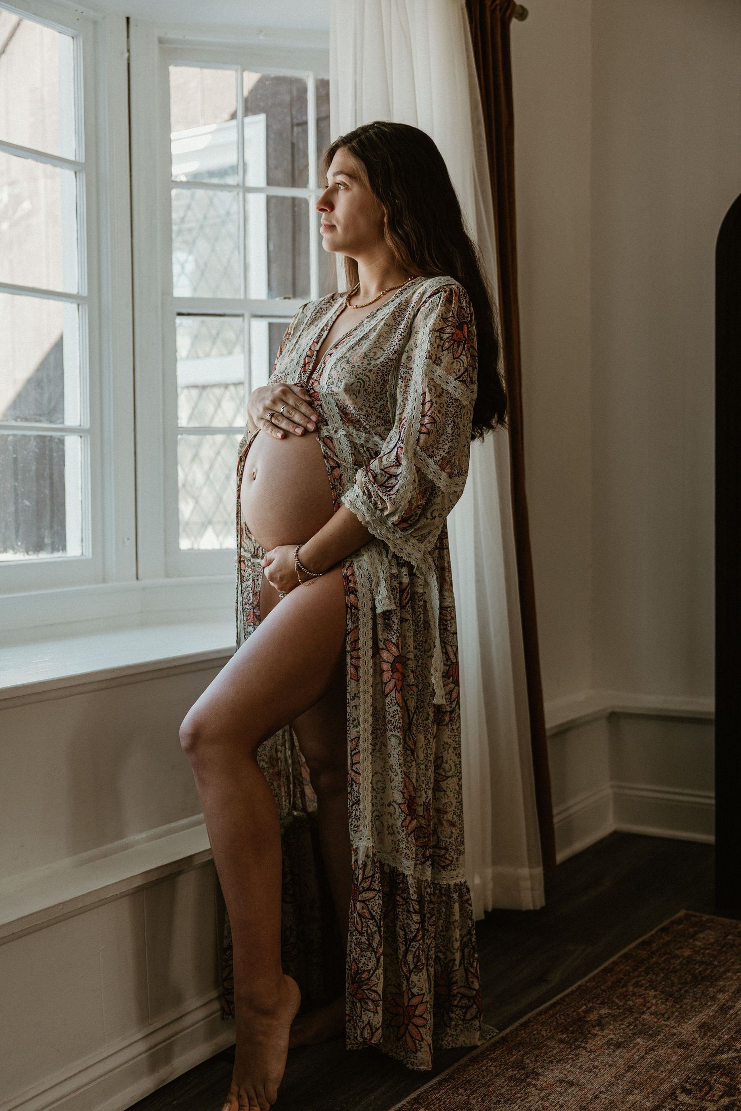 Elegant maternity photo with lace robe against window light and hardwood floors.
