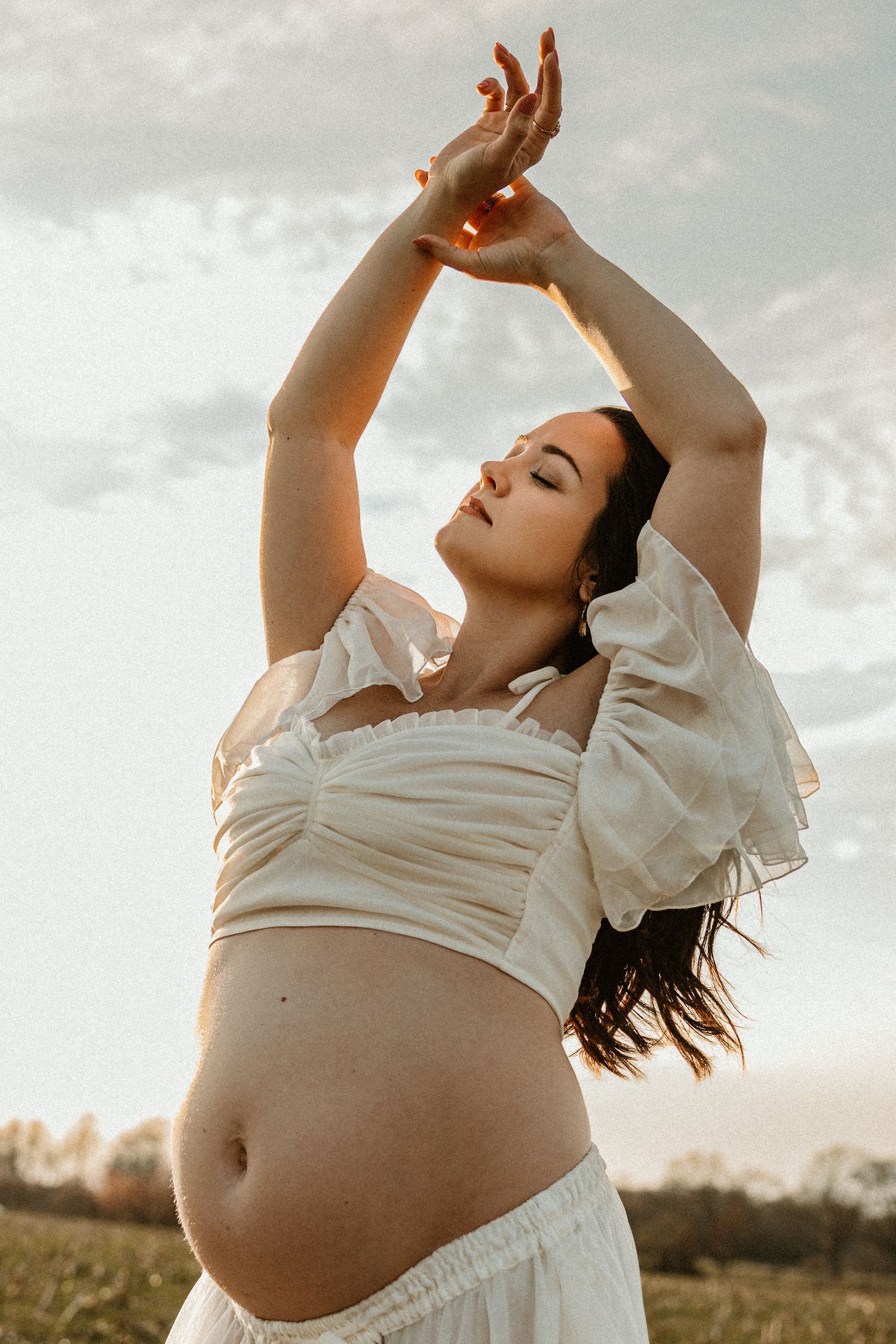 Pregnant woman in white crop top and skirt poses outdoors at golden hour with arms raised gracefully against cloudy sky.