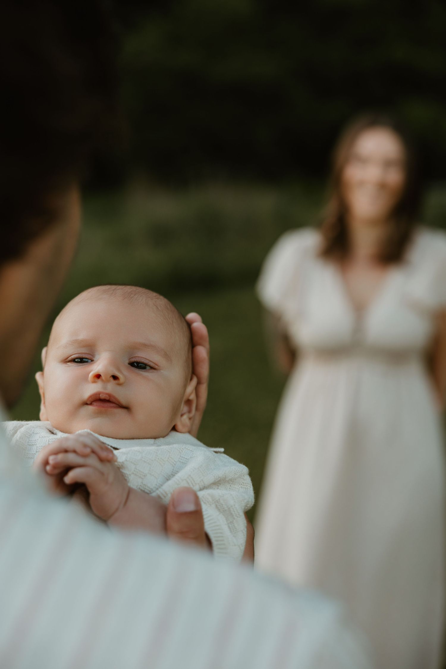 A newborn baby in white clothing being held up by a parent outdoors, with a smiling woman in a white dress in the background.