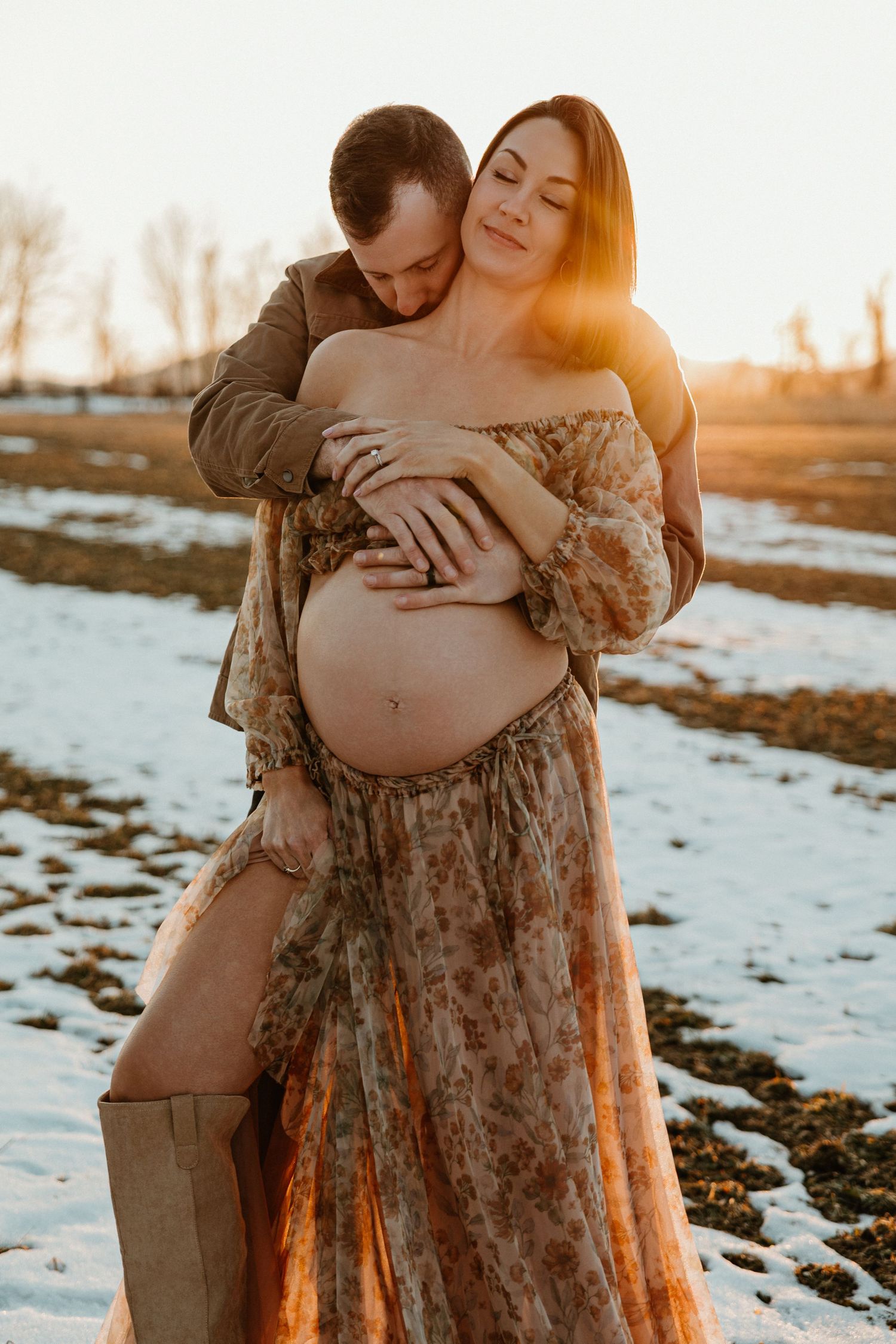 A couple embraces in a snowy field at sunset while wearing a floral dress and military uniform.