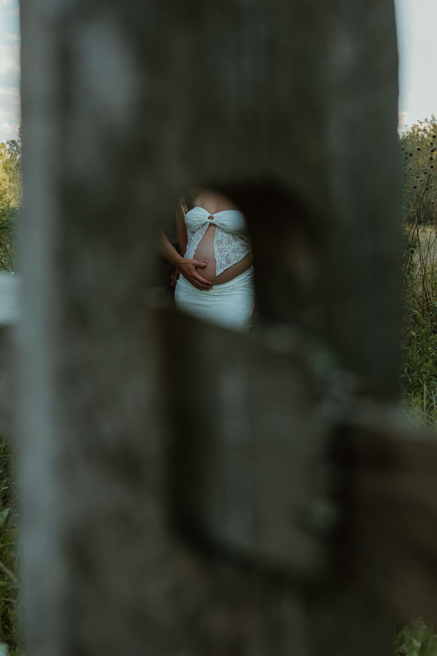 Pregnant woman in white lace dress cradling baby bump, photographed through rustic wooden gap outdoors.