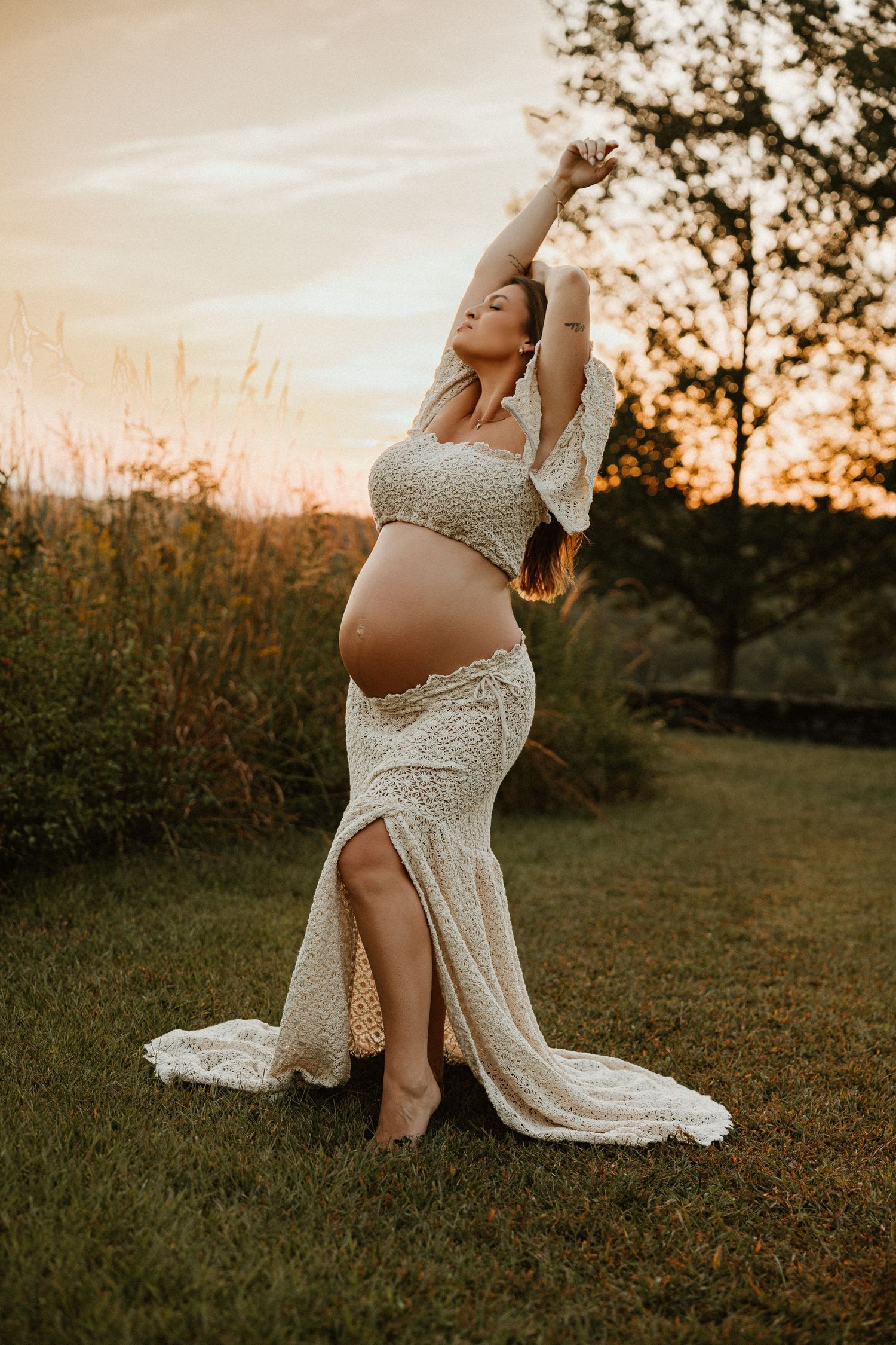 Expectant mother in flowing white lace dress poses gracefully during golden hour outdoor photoshoot.