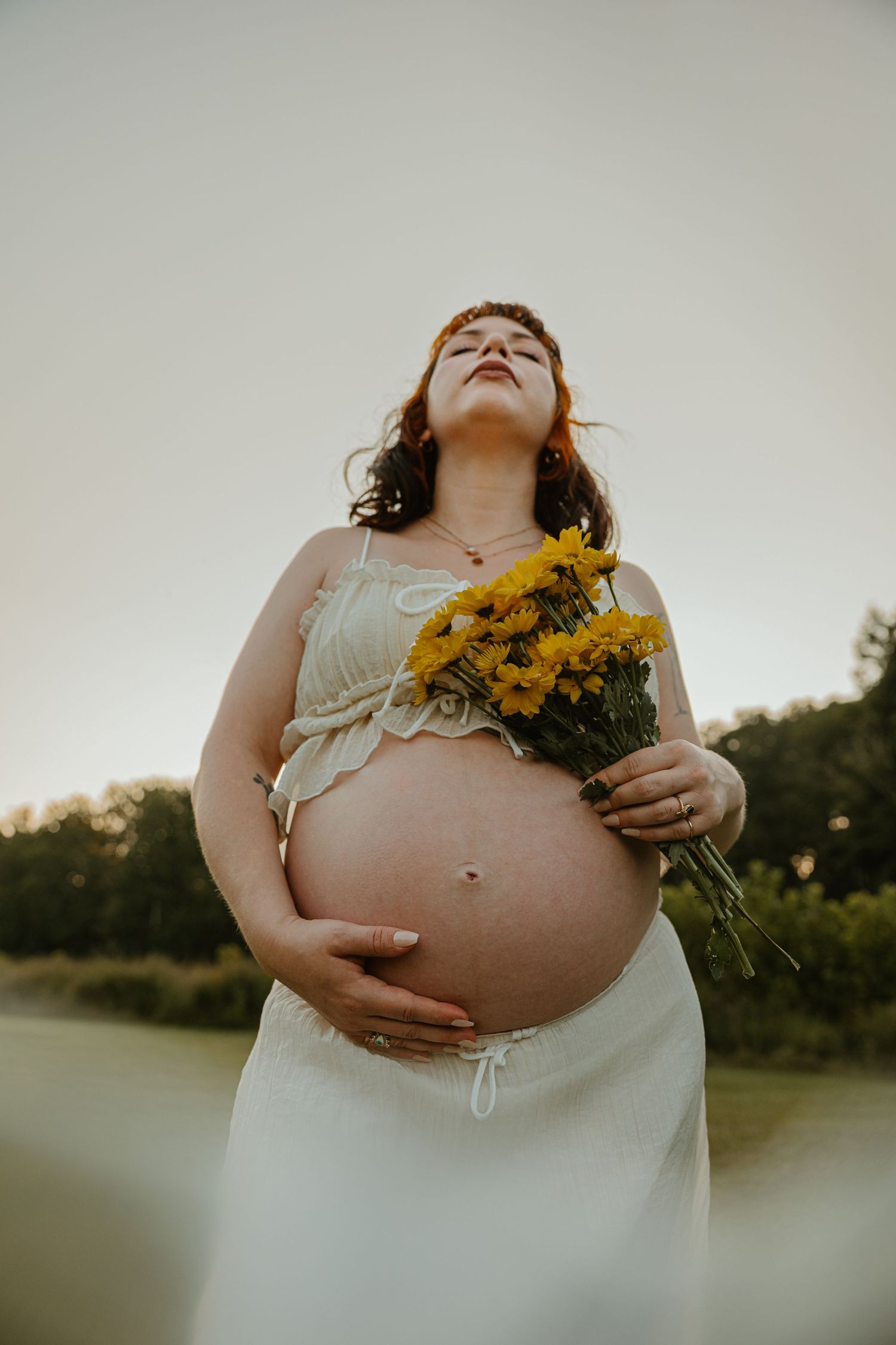 Pregnant woman in white dress holding yellow flowers outdoors, cradling her bare baby bump near a serene lakeside.