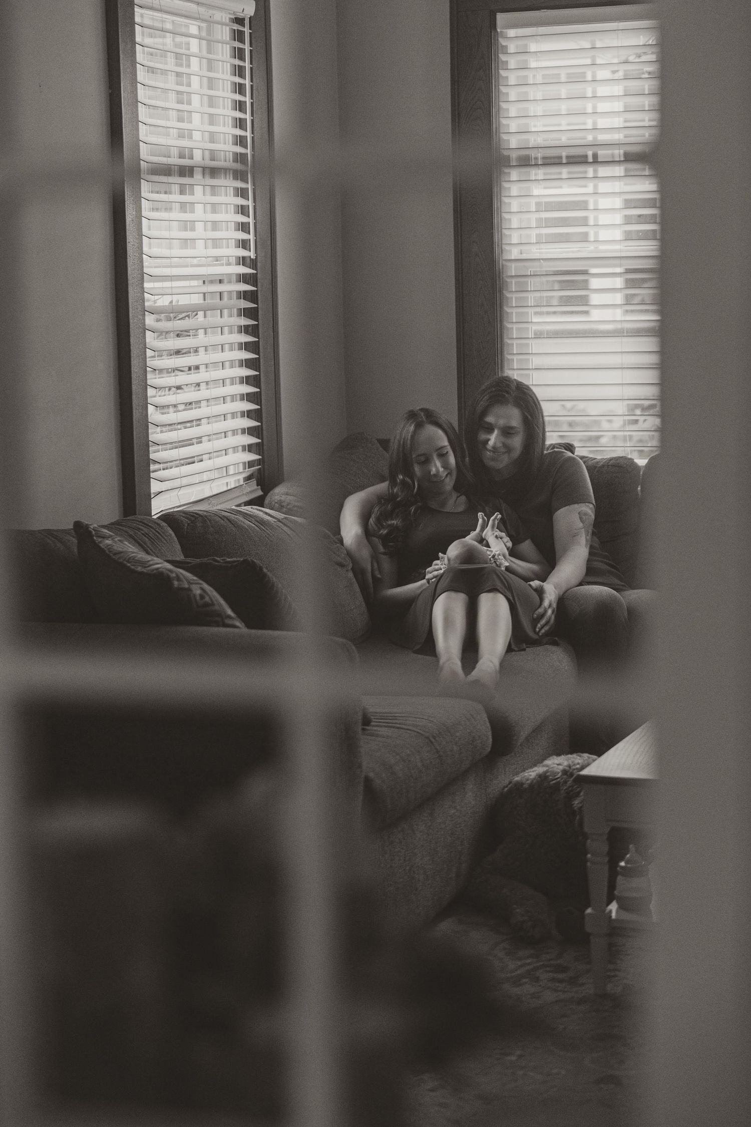 Black and white photo of two women sitting together on a couch by windows, sharing a warm moment.