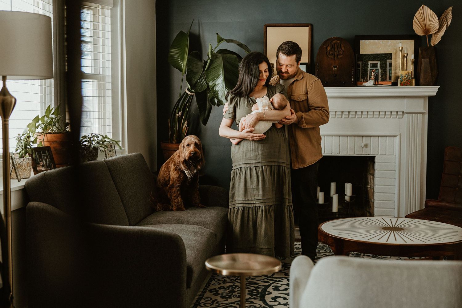 A couple stands in a cozy living room holding their newborn baby, with a dog and fireplace in the background.