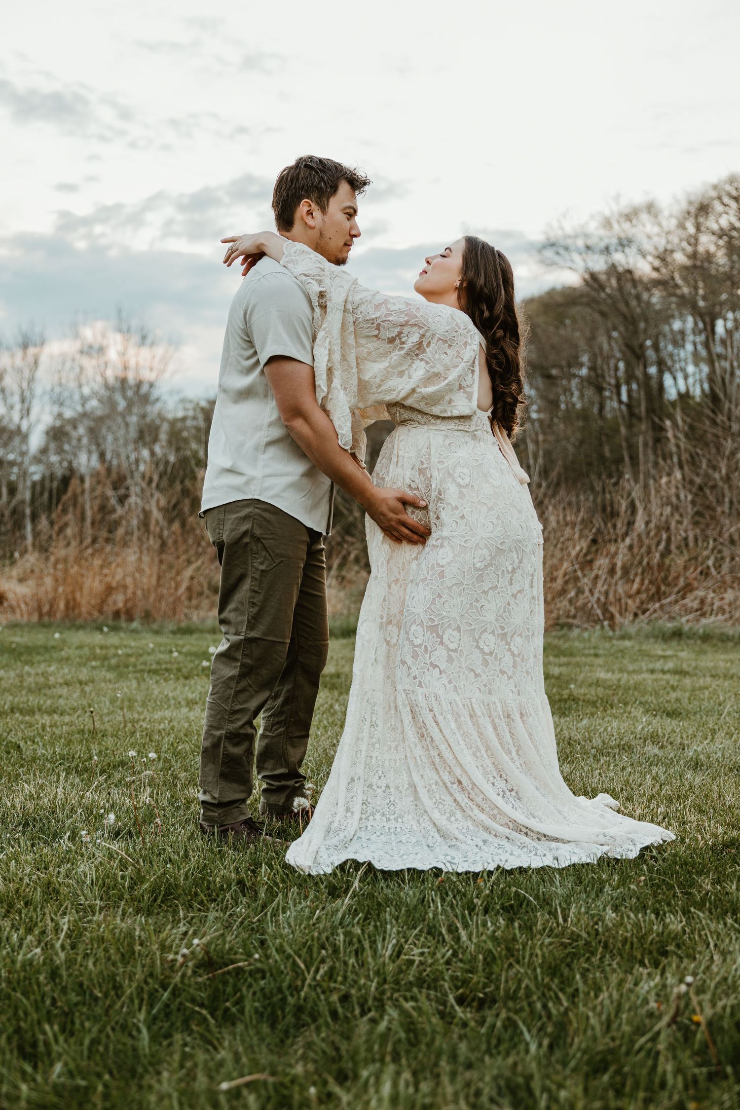 A couple shares a romantic moment outdoors, woman in white lace maternity gown embracing man in olive shirt on green grass.