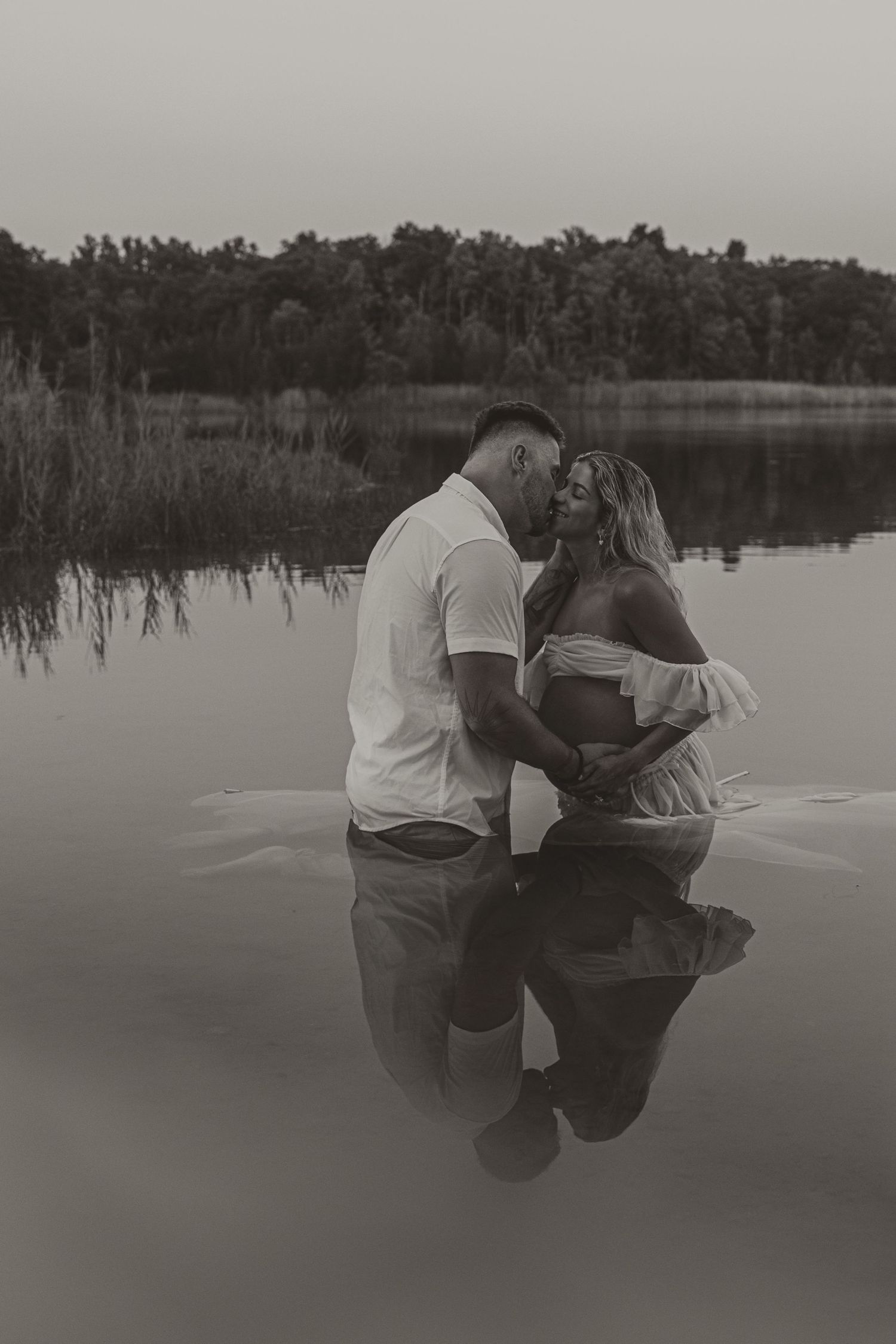 Black and white photo of a couple standing in a lake, the woman visibly pregnant, sharing a tender moment.