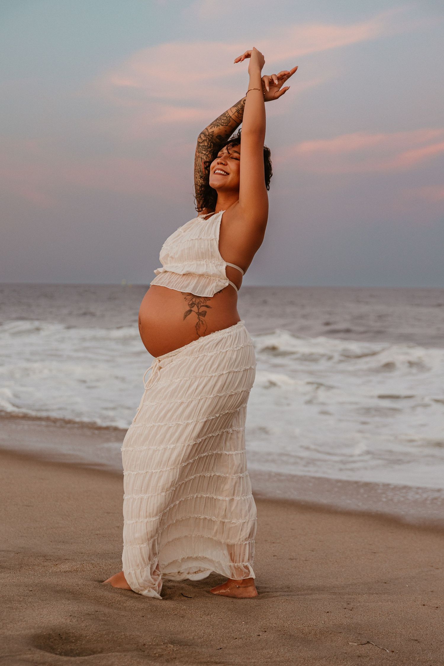 Maternity beach photoshoot at sunset in flowing white dress.
