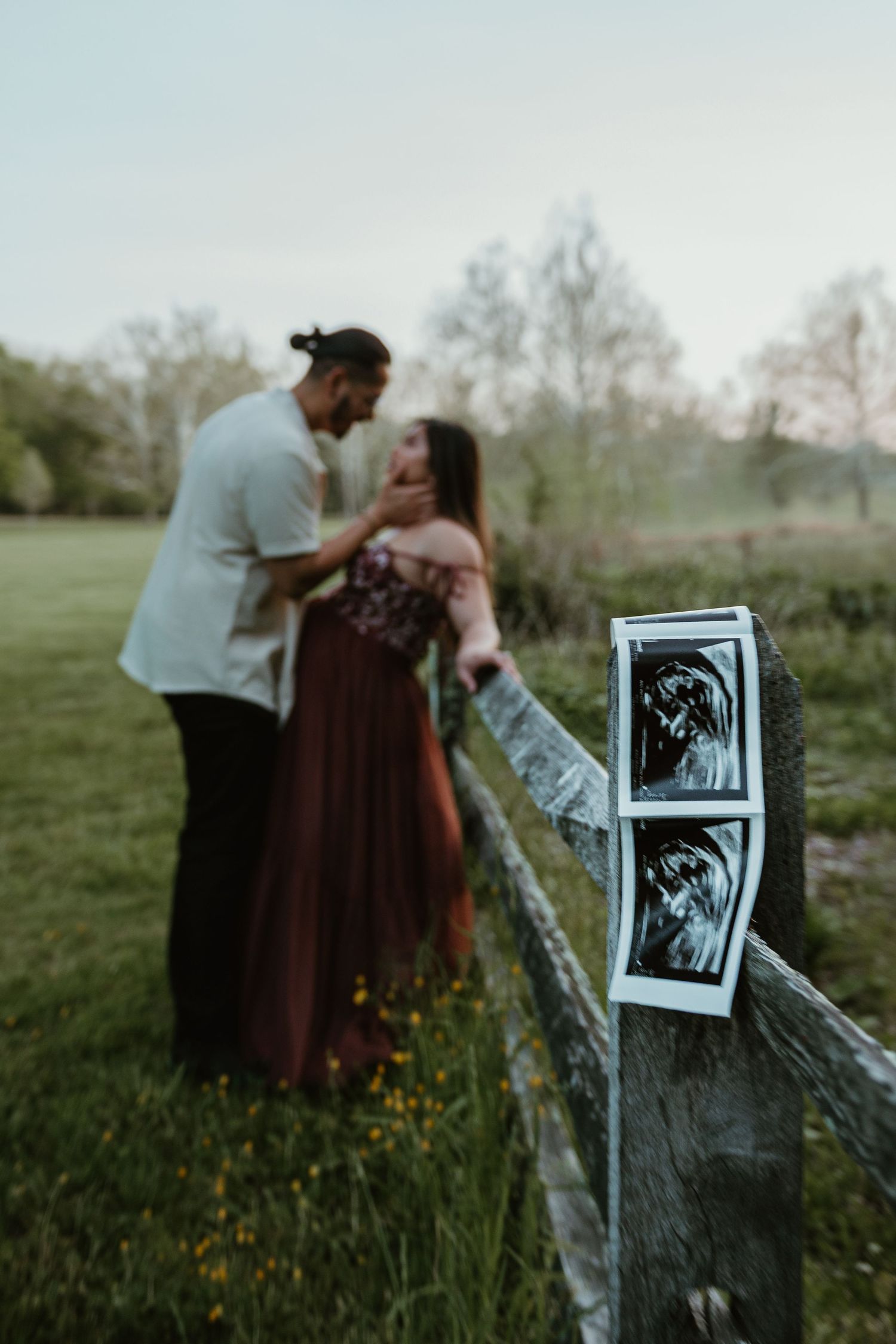 Couple sharing a romantic moment by a rustic fence with ultrasound photos in the foreground, pregnancy announcement.