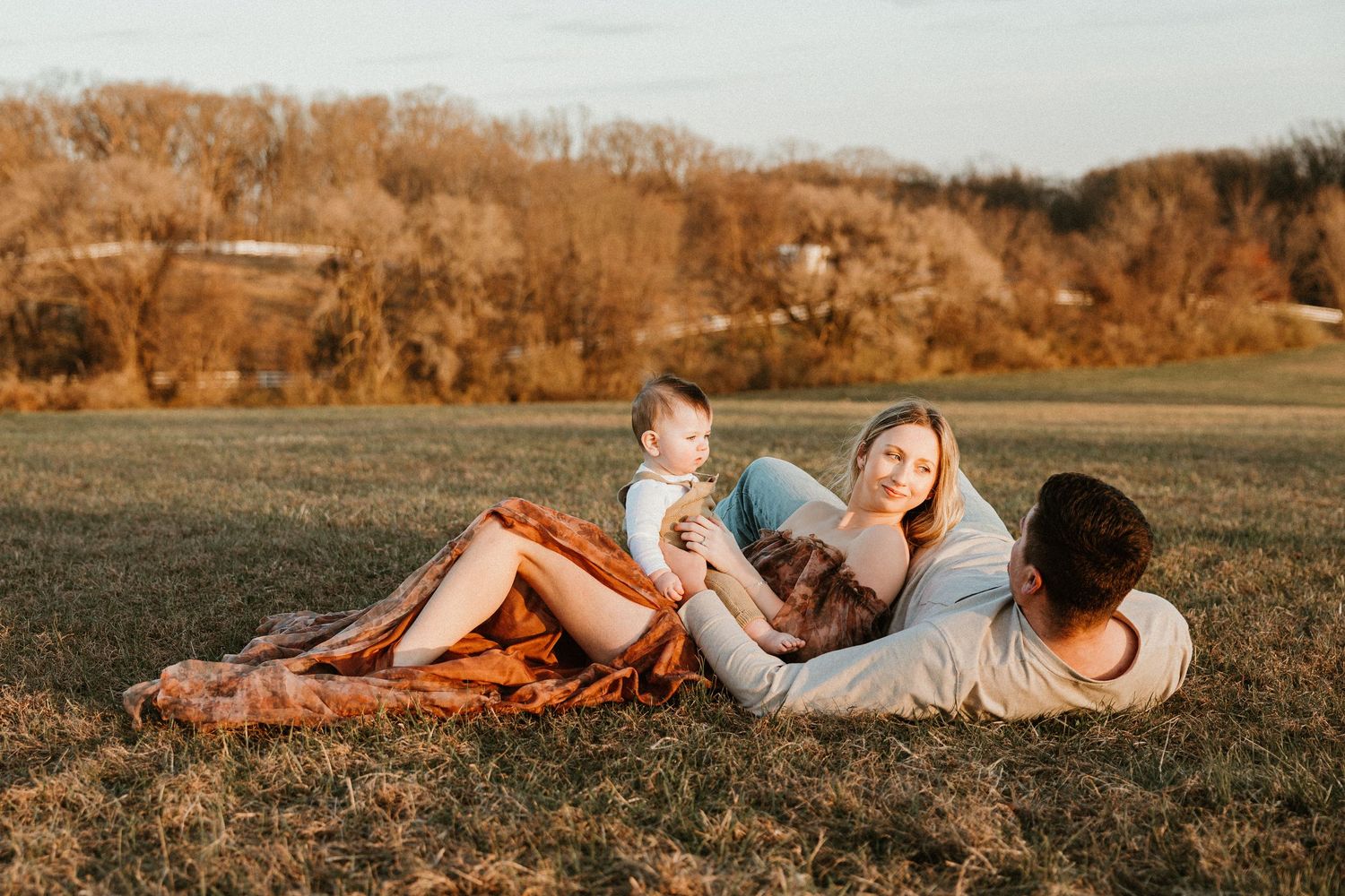 Family relaxing on a blanket in a sunny autumn field, parents and baby enjoying a golden hour outdoor moment.