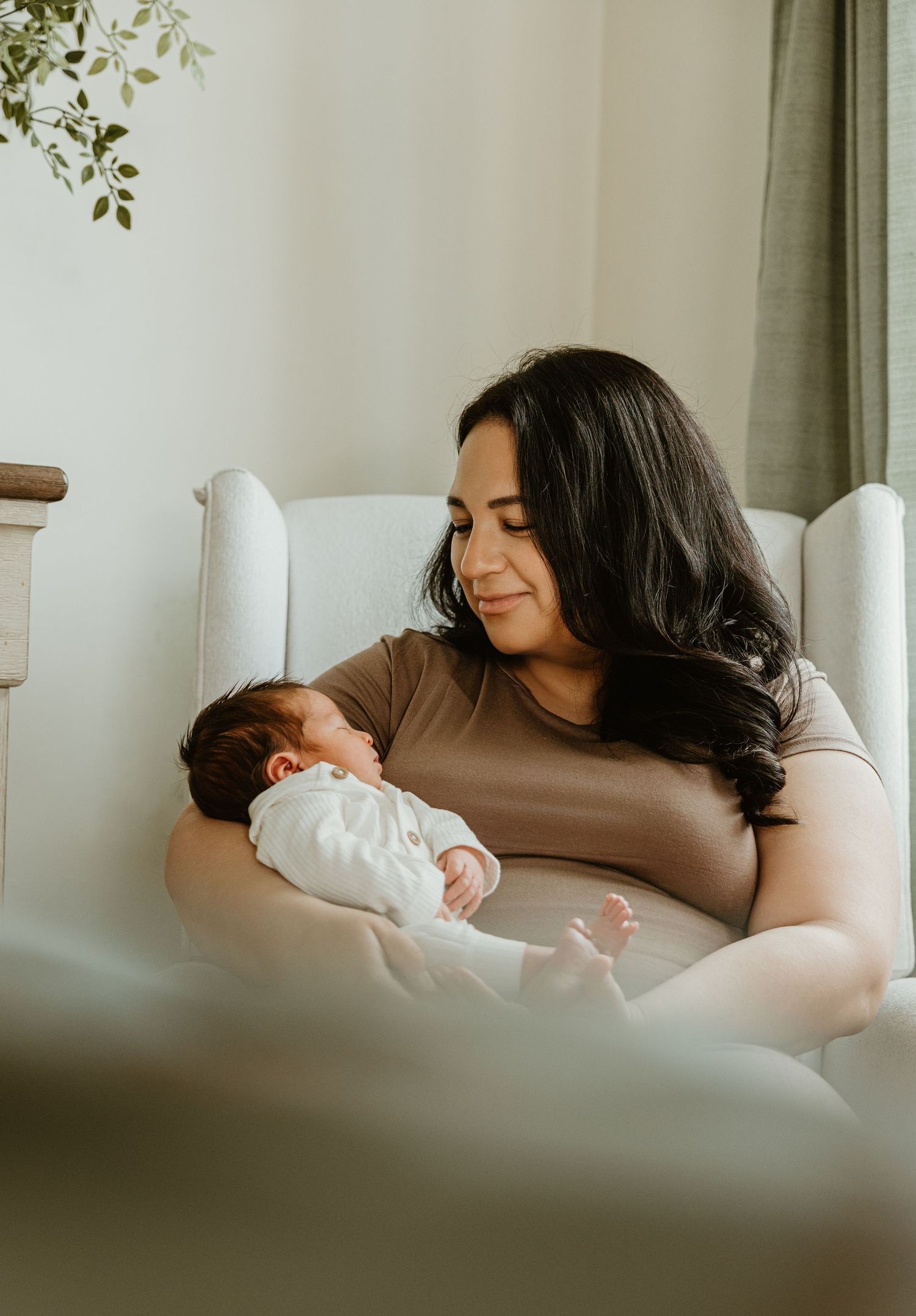 A mother with dark hair sits in a white chair, lovingly gazing at her newborn baby dressed in white.
