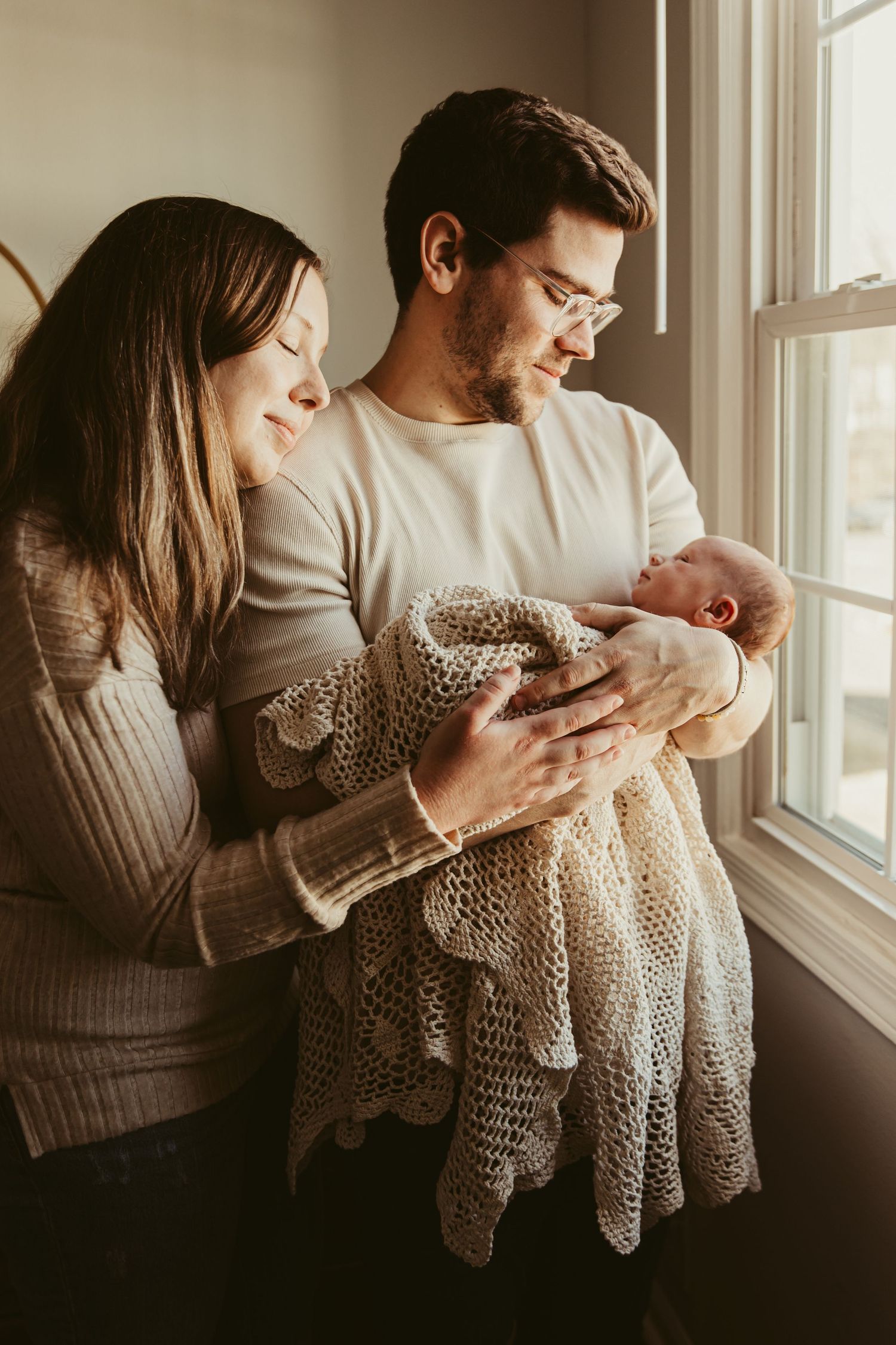Couple in cozy sweaters holding their newborn baby wrapped in knit blanket by window.