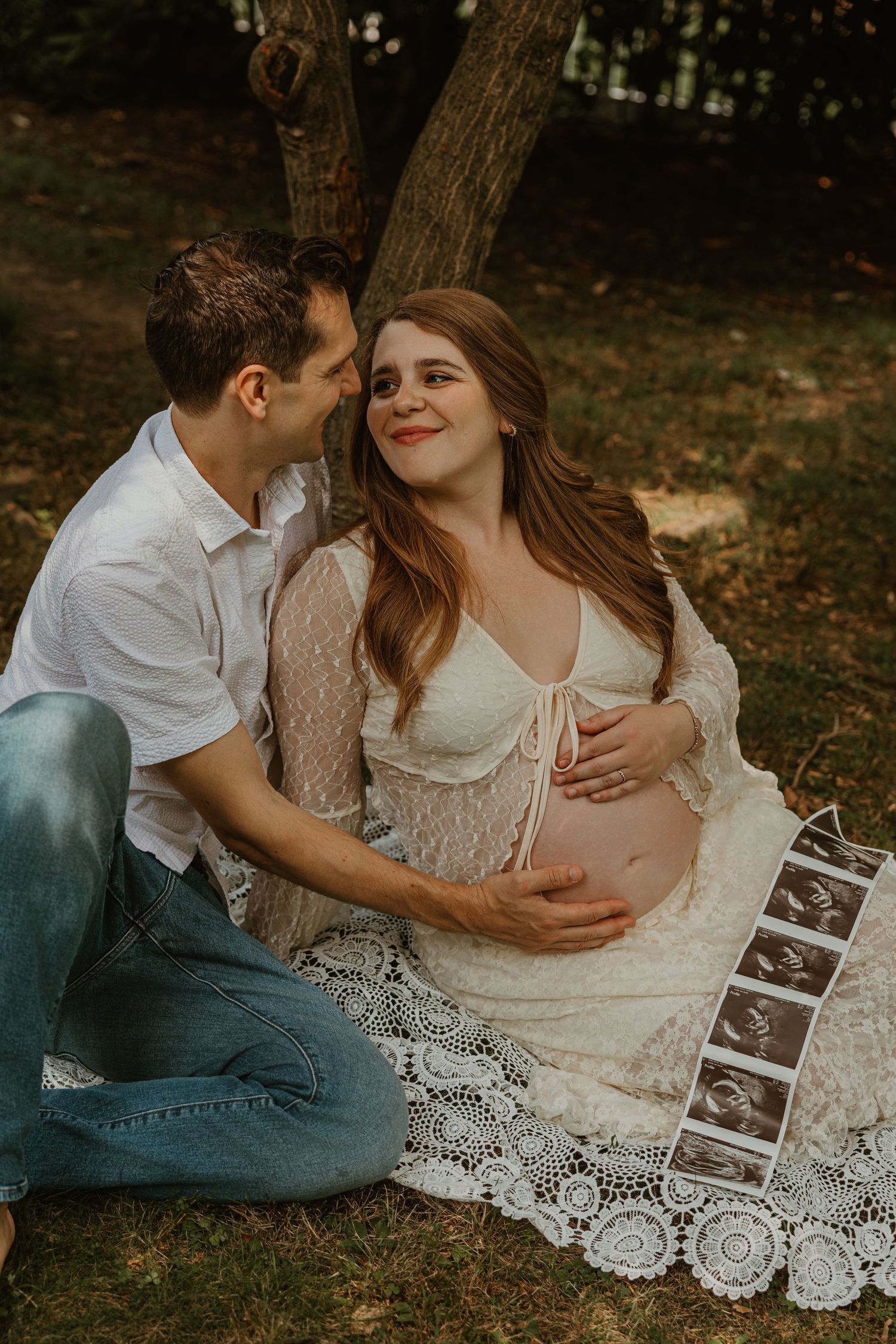 Pregnant couple sitting outdoors on lace blanket, man whispering to smiling woman holding baby bump with ultrasound photos nearby.