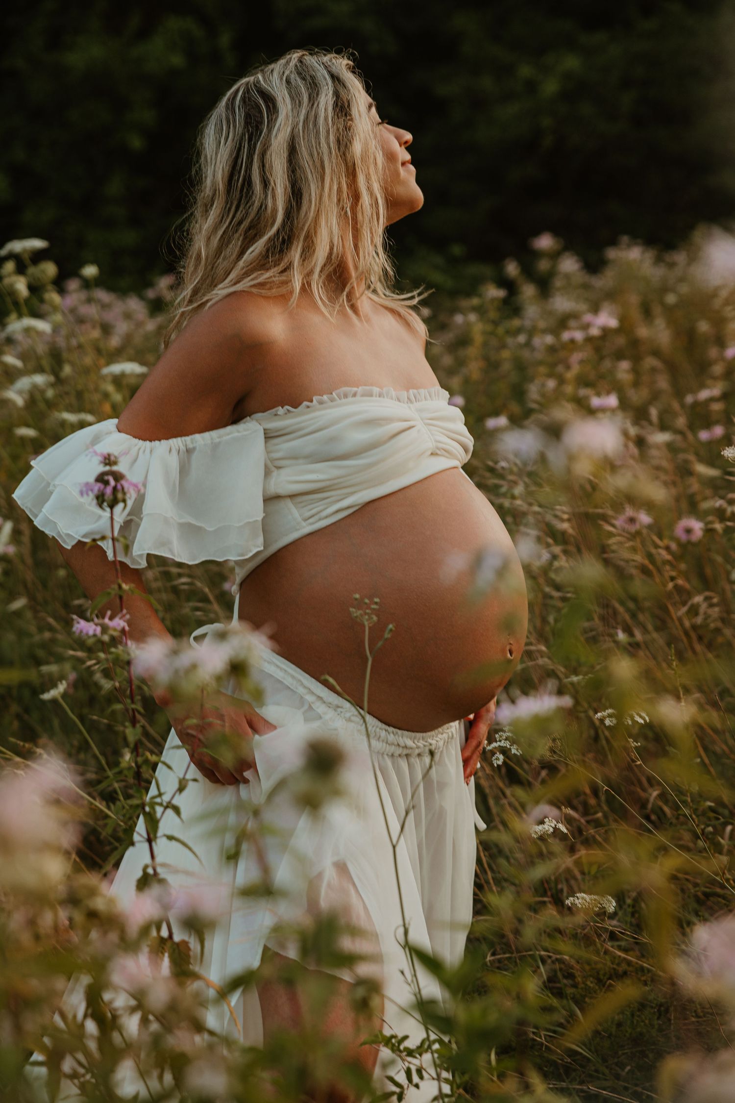 Pregnant woman in white off-shoulder dress standing in wildflower field, cradling baby bump.