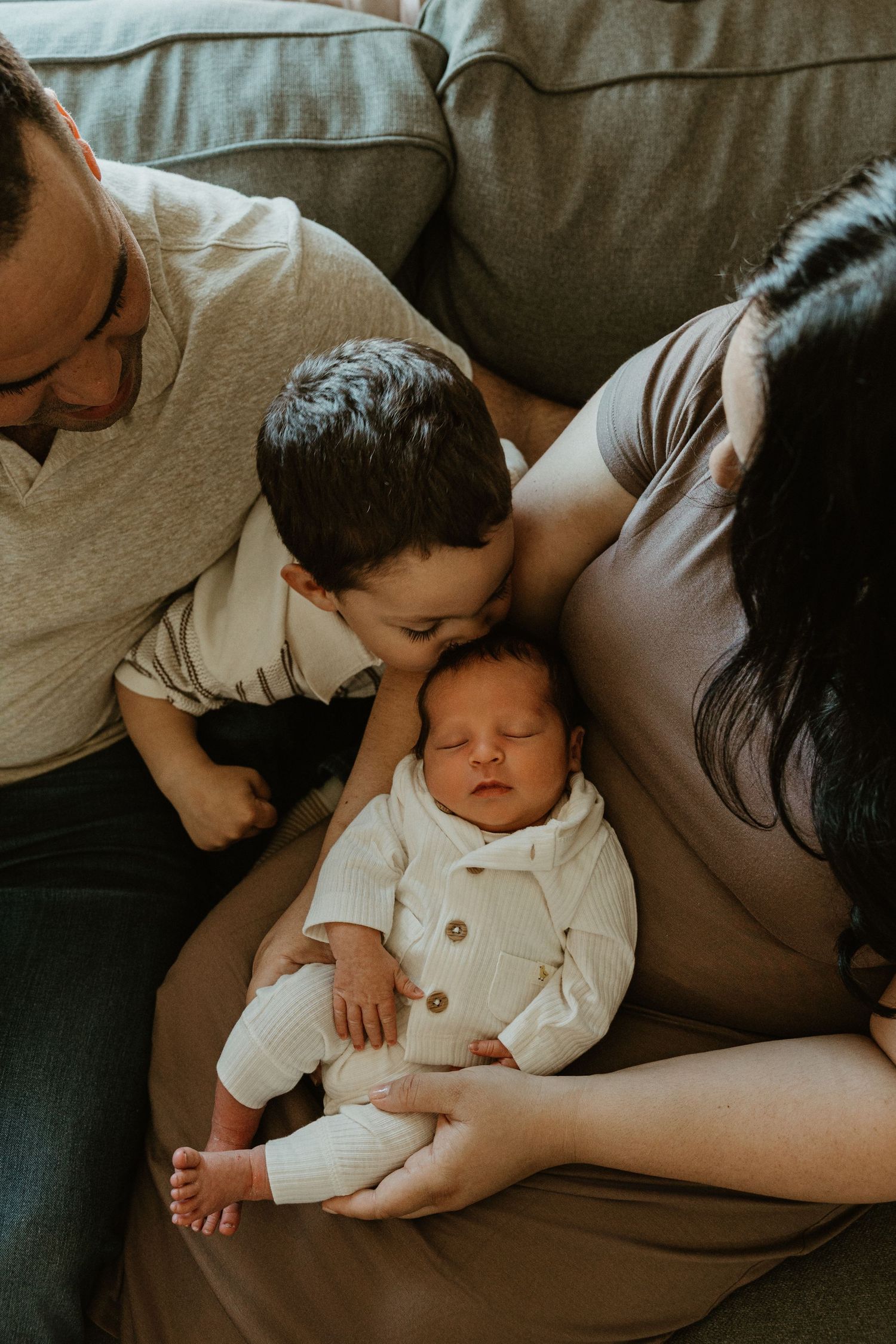 Family with newborn baby, older sibling giving a gentle kiss while parents look on lovingly on a cozy couch.