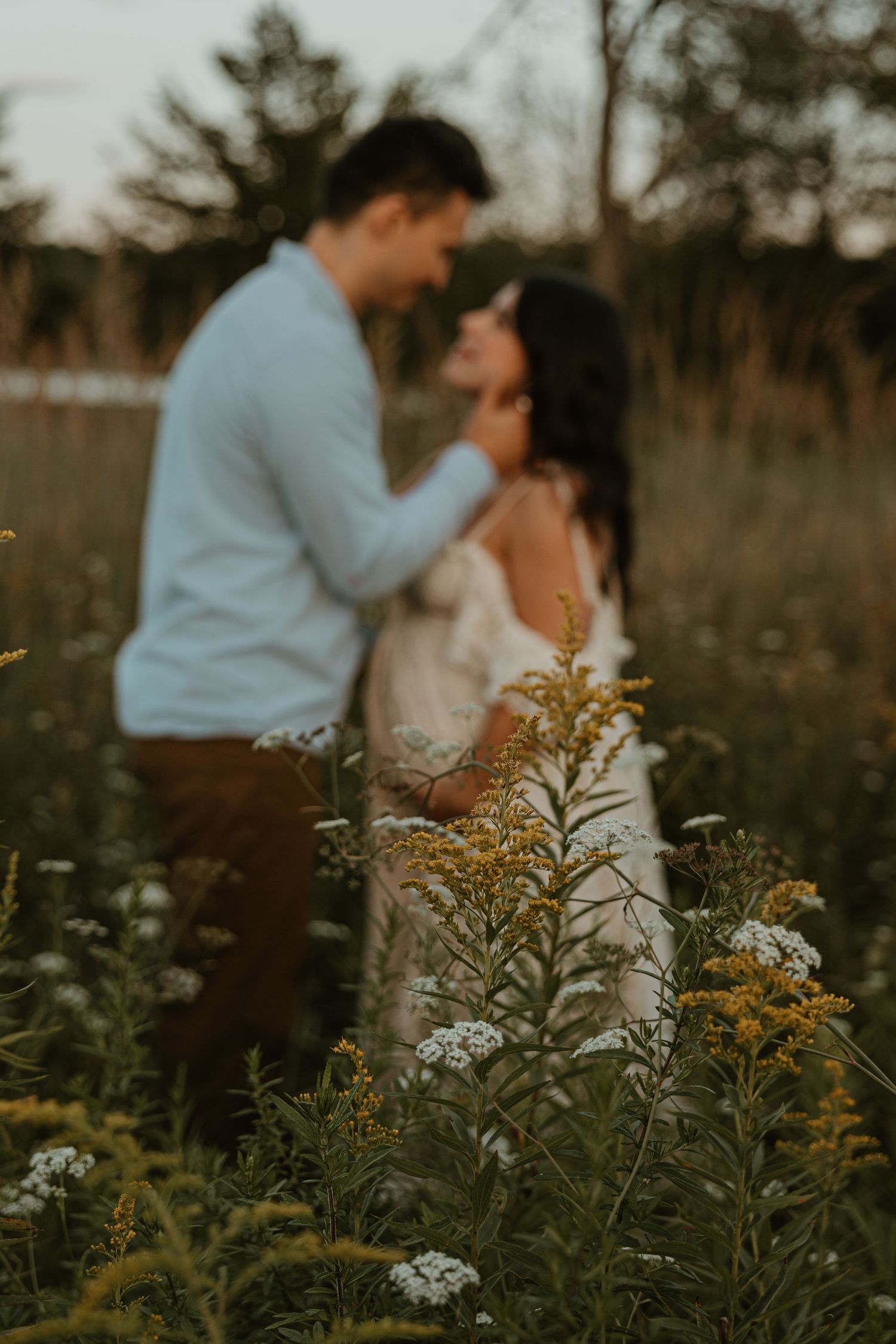 Couple embracing in a wildflower meadow at golden hour, with yellow and white blooms in soft focus foreground.