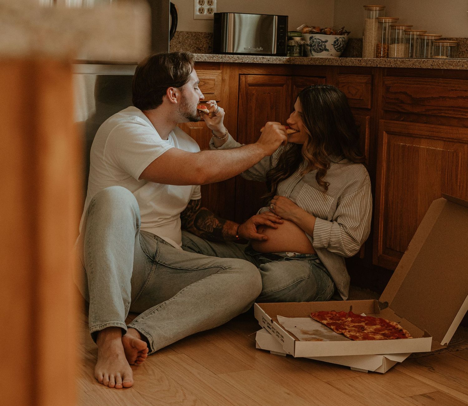 Couple sitting on kitchen floor sharing pizza, woman appears pregnant, enjoying a casual romantic moment together.