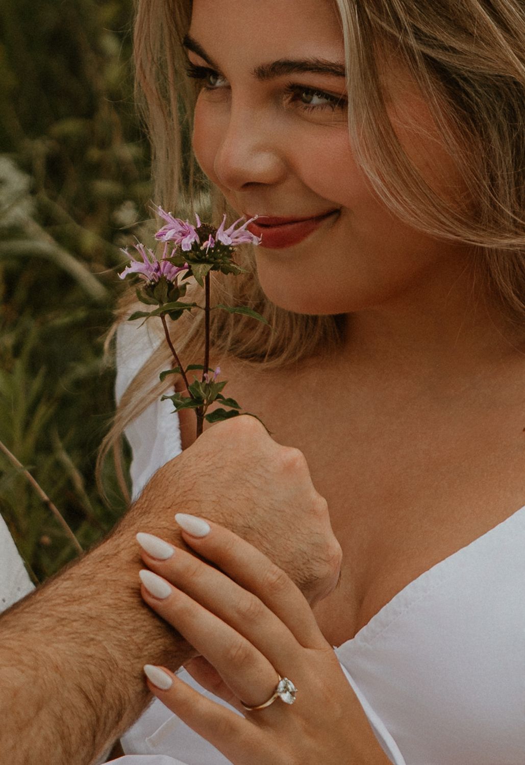 A smiling woman with blonde hair holds pink wildflowers, showcasing a diamond engagement ring on her hand.