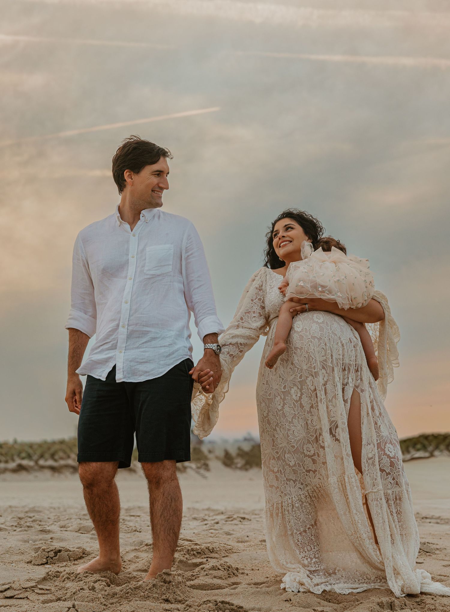 Couple holding hands on beach at sunset, man in white shirt, pregnant woman in lace maternity gown.