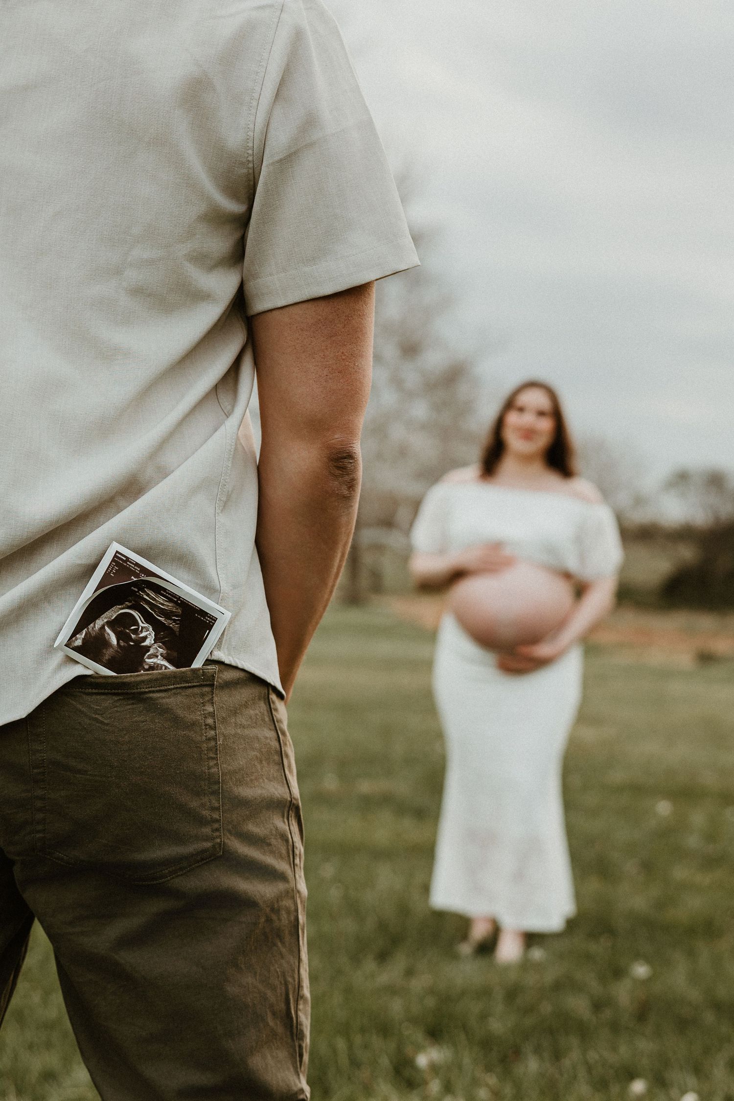 Man holding ultrasound photo in back pocket while pregnant woman in white dress stands in field.