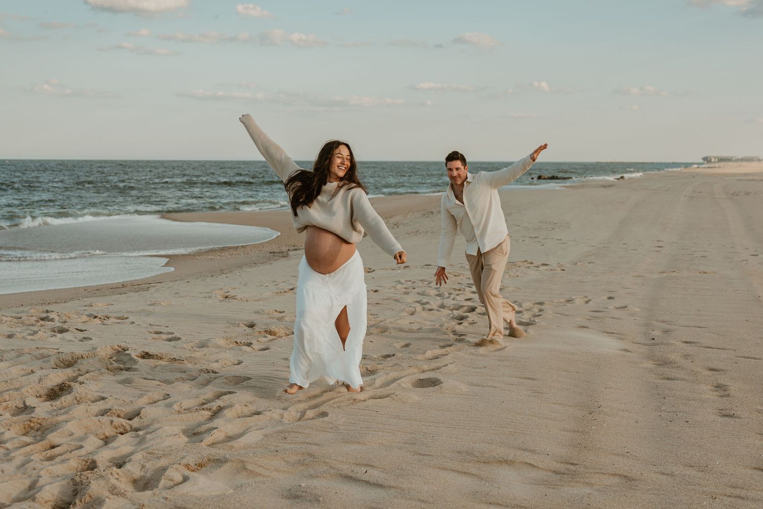 Pregnant woman in white dress carried by man on sandy beach, joyful maternity photo shoot by the ocean.