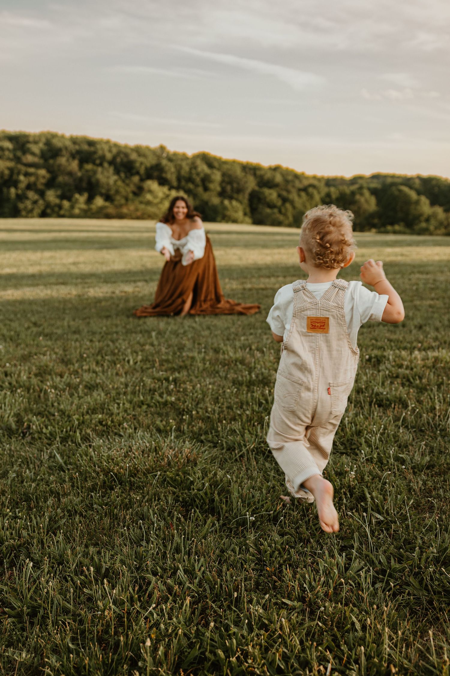 Toddler in overalls running barefoot toward pregnant mother sitting in green field at sunset.