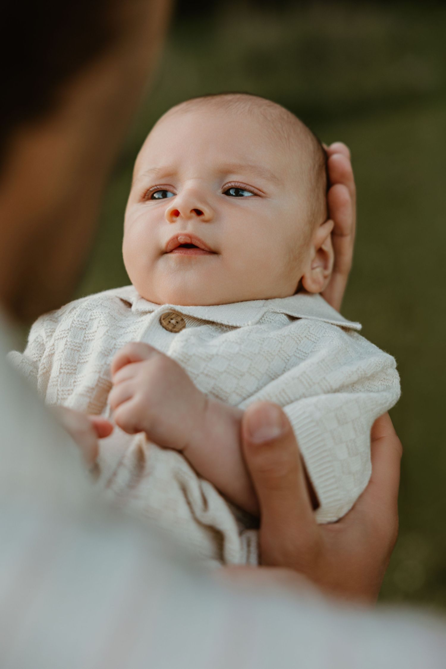 A newborn baby in a cream knit outfit being cradled in adult hands, gazing upward with curious eyes.