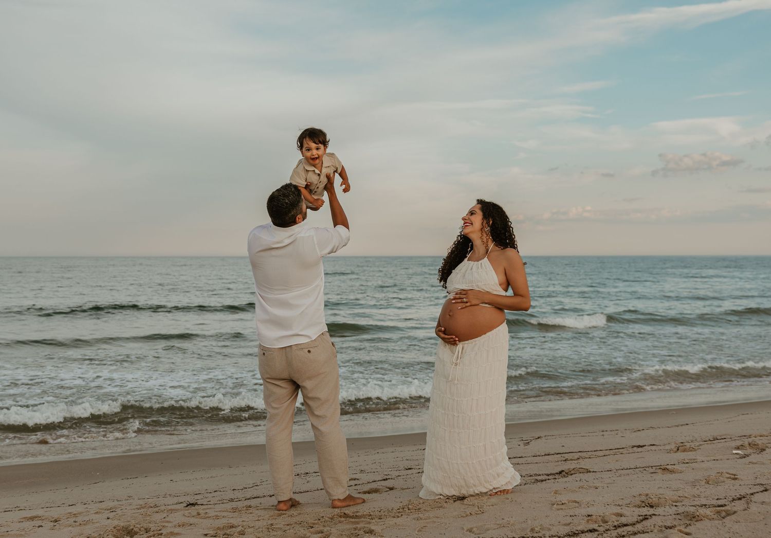 Pregnant woman in white dress with family on beach, husband lifting toddler, ocean backdrop at sunset.