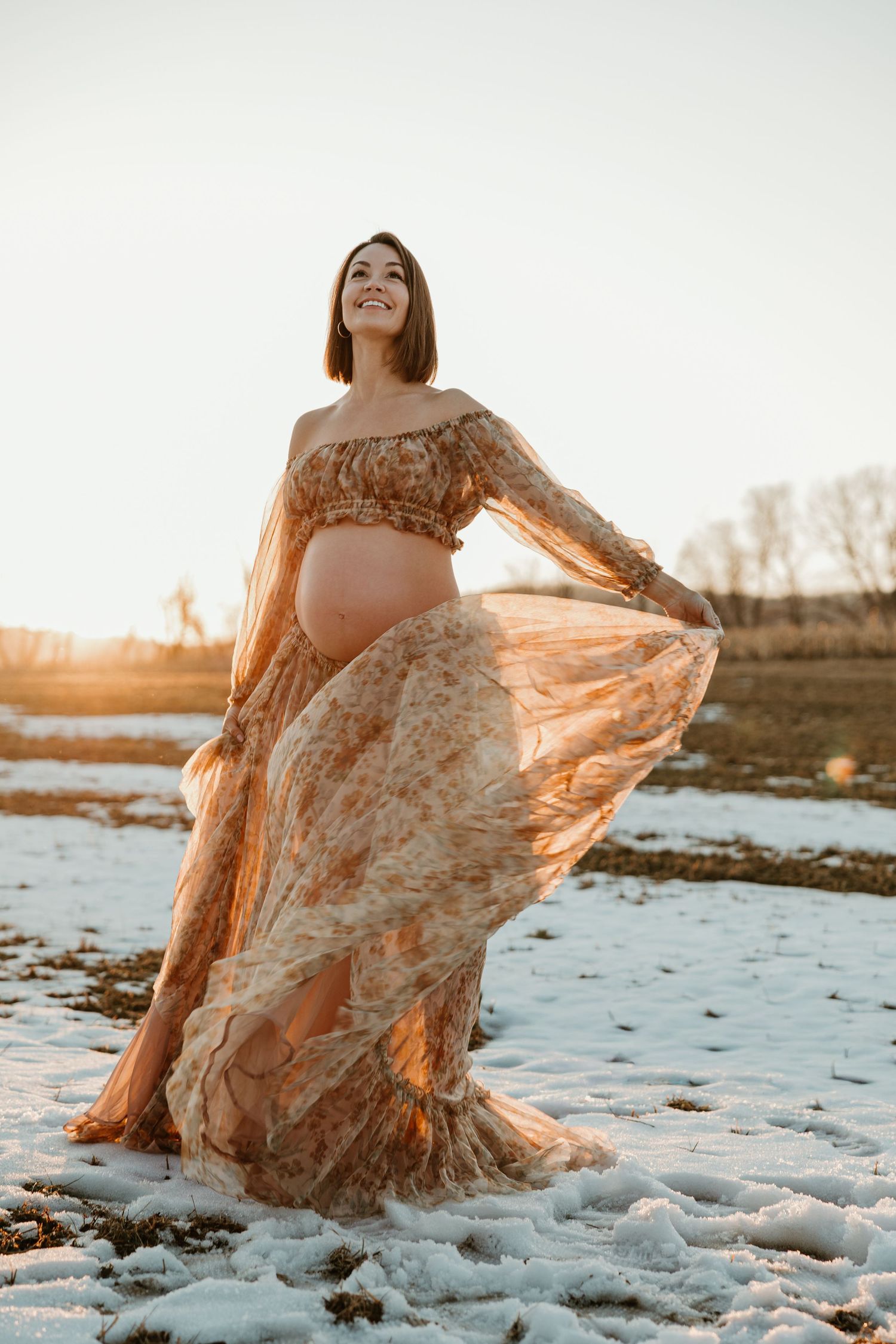 Maternity photo session at sunset with flowy floral dress in snow covered landscape showing movement and golden light.