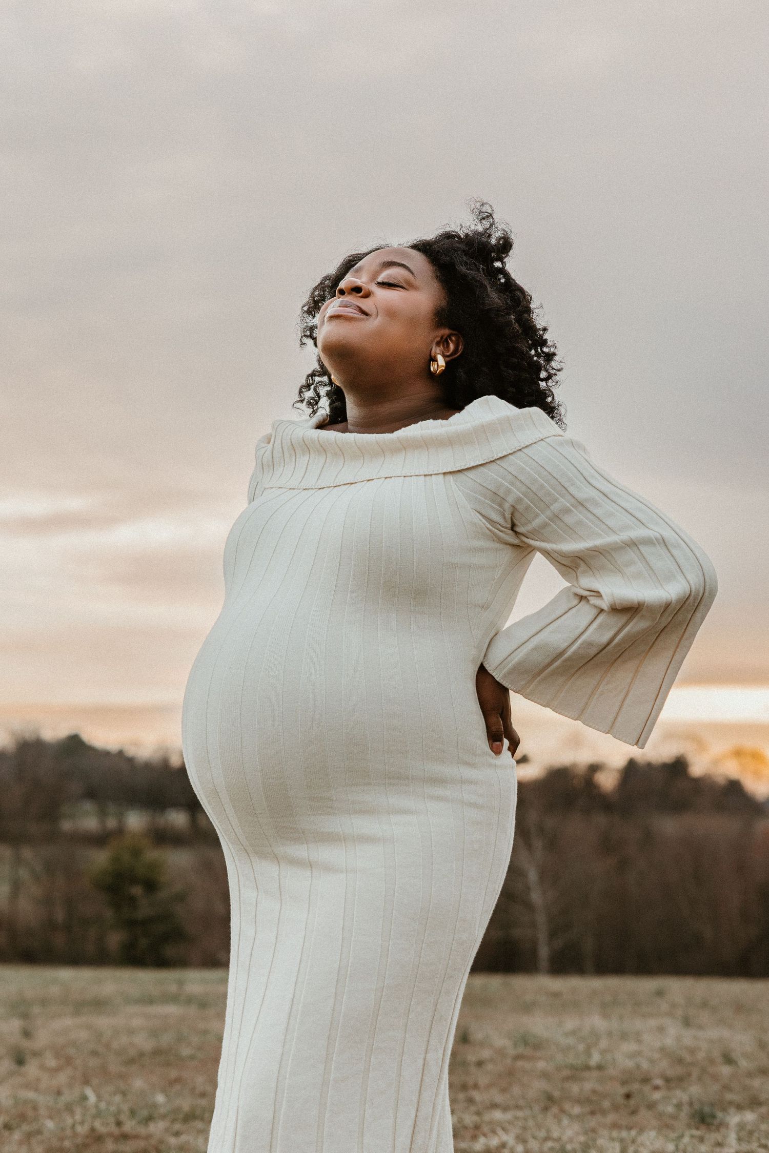 Pregnant woman in white ribbed dress poses outdoors at sunset, smiling with eyes closed, hand on hip.