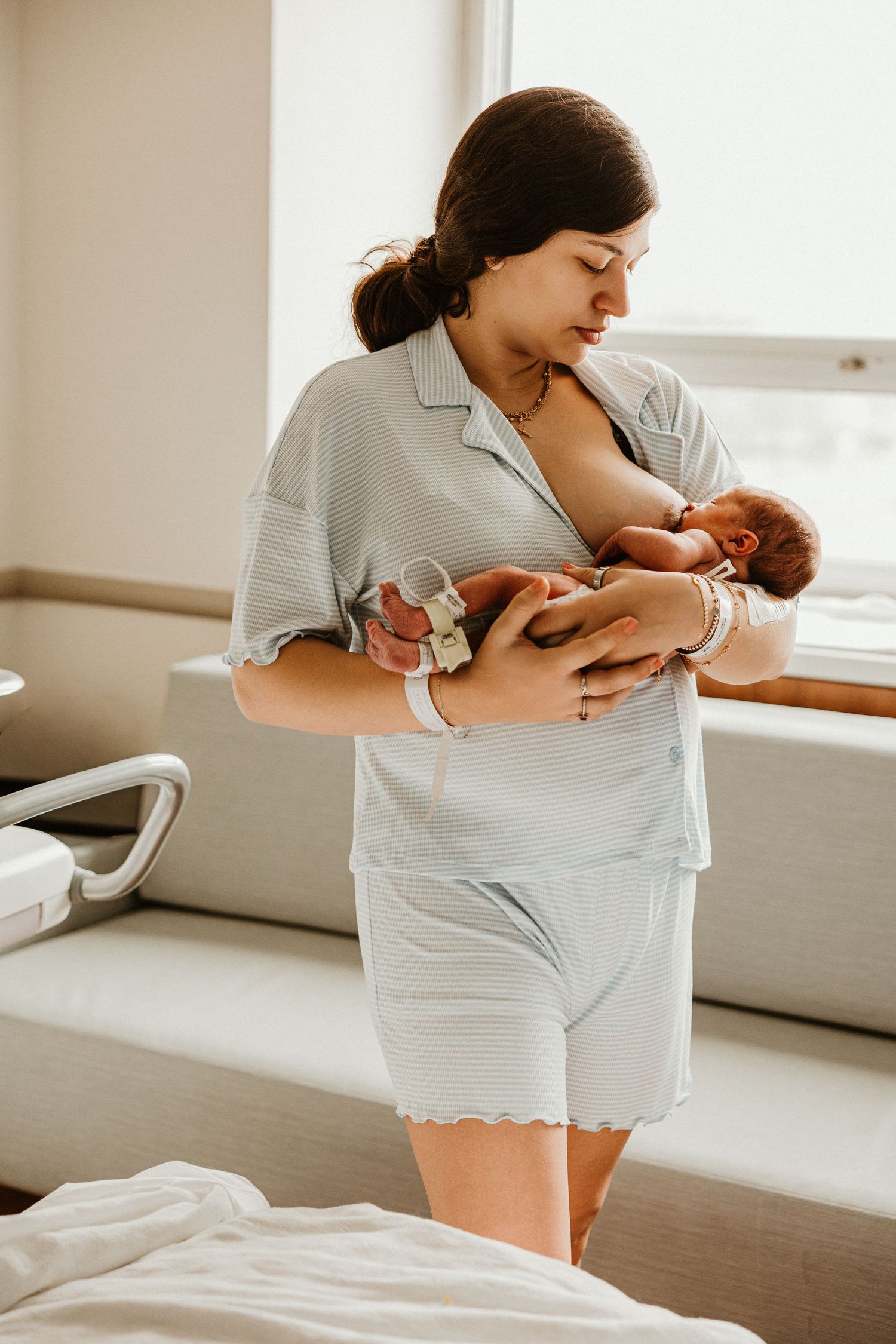 New mother in light blue pajamas breastfeeding her newborn baby while standing in a hospital room.