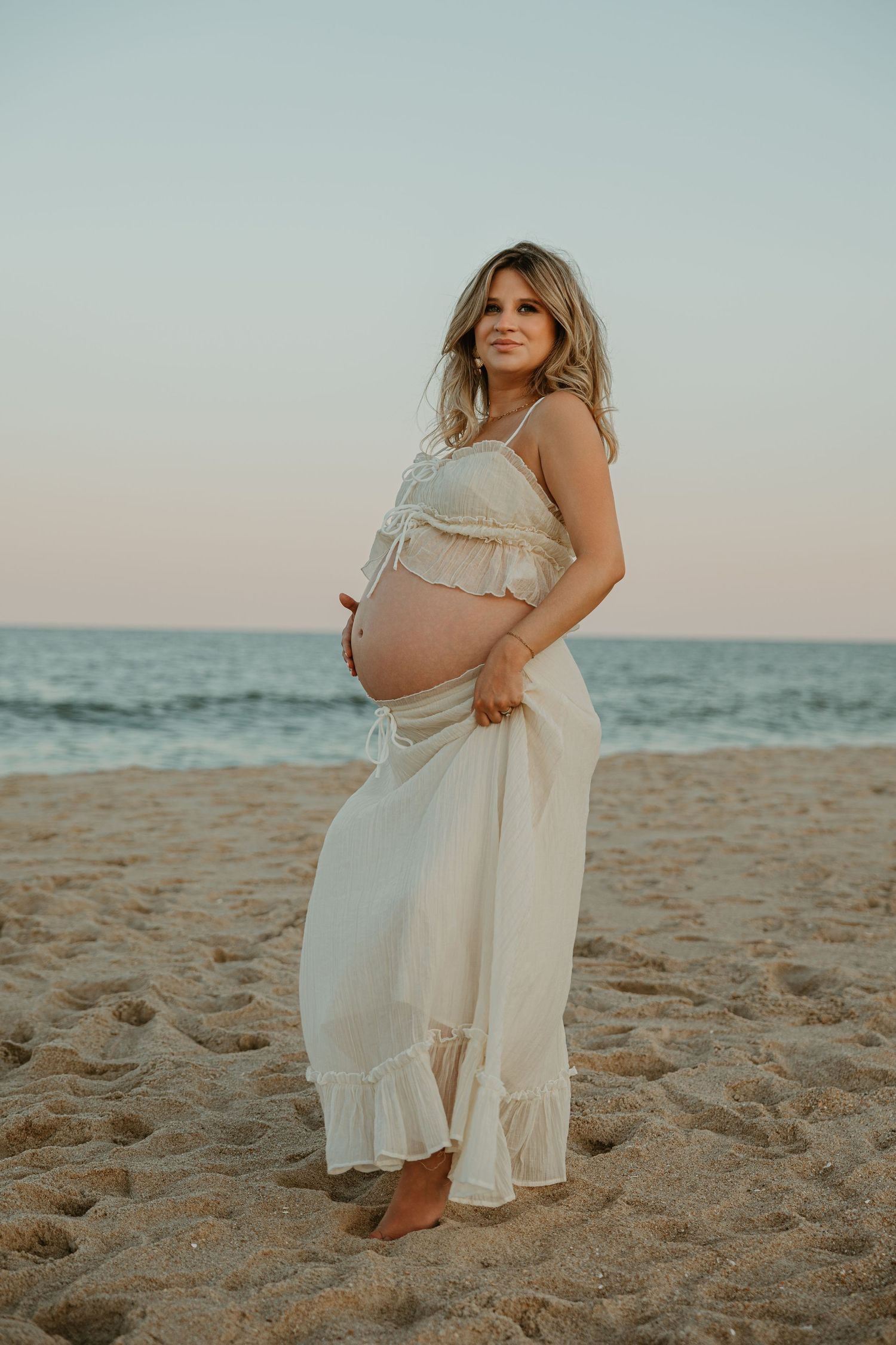 Pregnant woman in white crop top and maxi skirt posing on sandy beach at sunset with ocean in background.