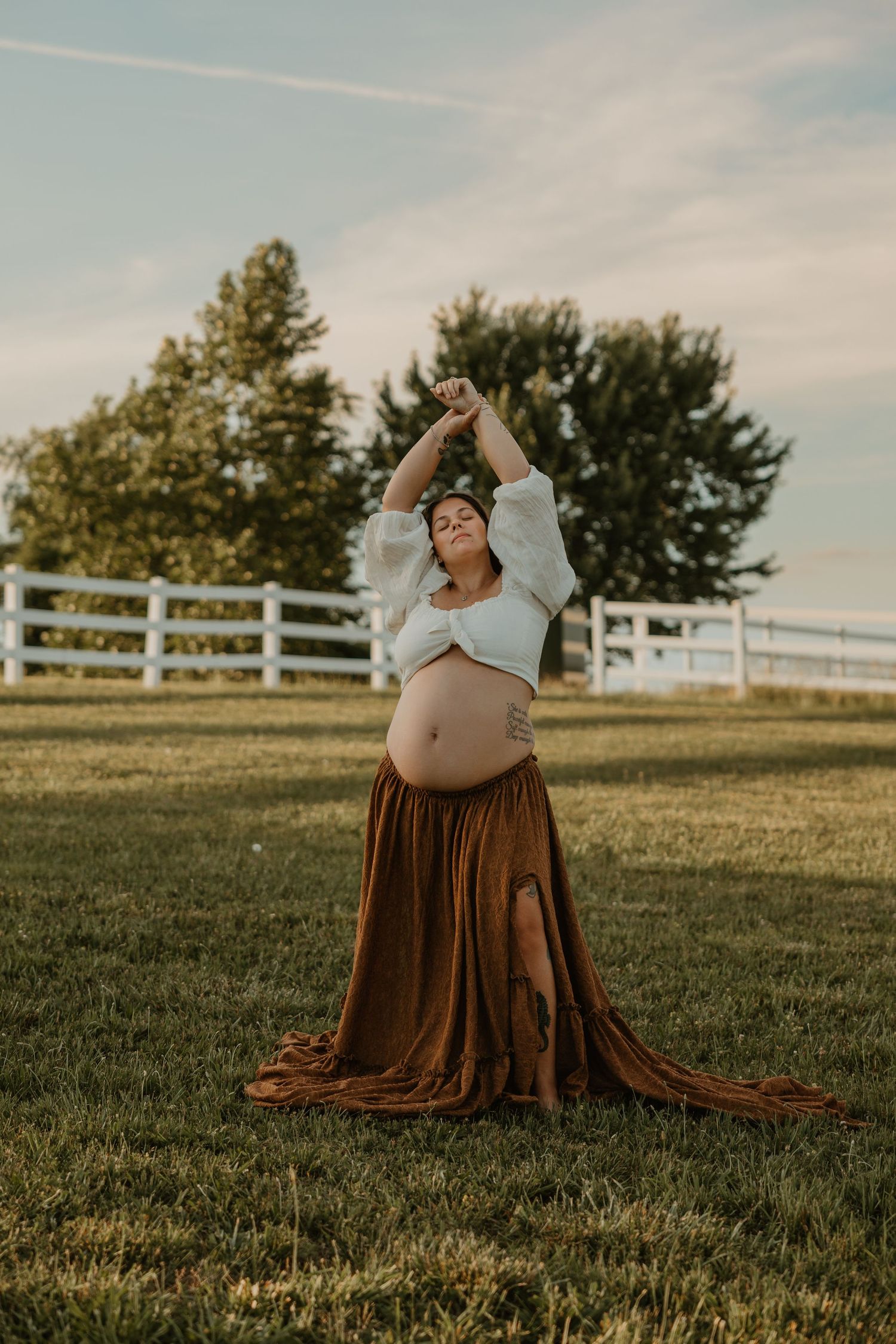 Pregnant woman in white crop top and brown fringe skirt posing outdoors on green grass with arms raised.