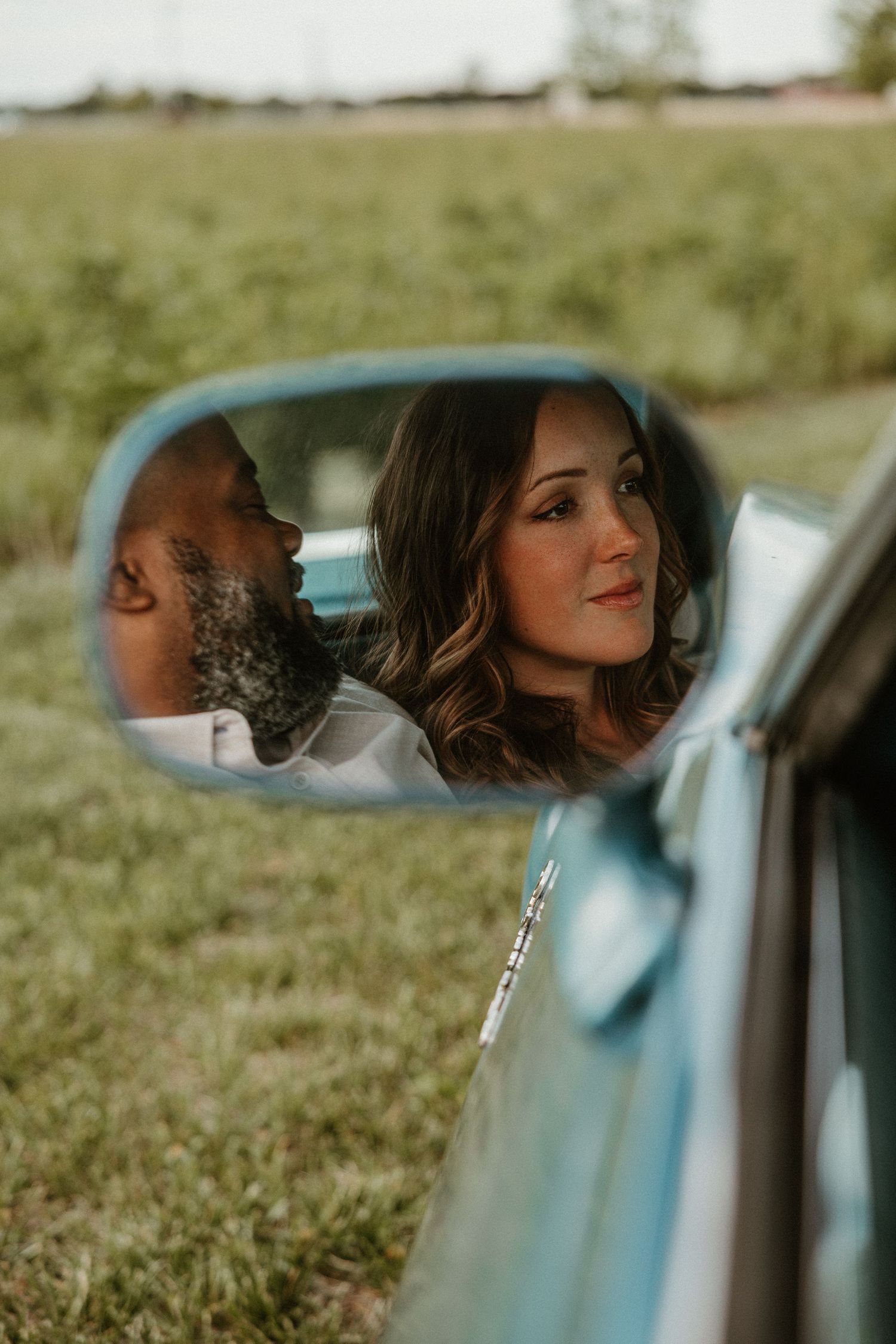 Couple reflected in vintage car side mirror, woman smiling softly, man beside her, green field background.