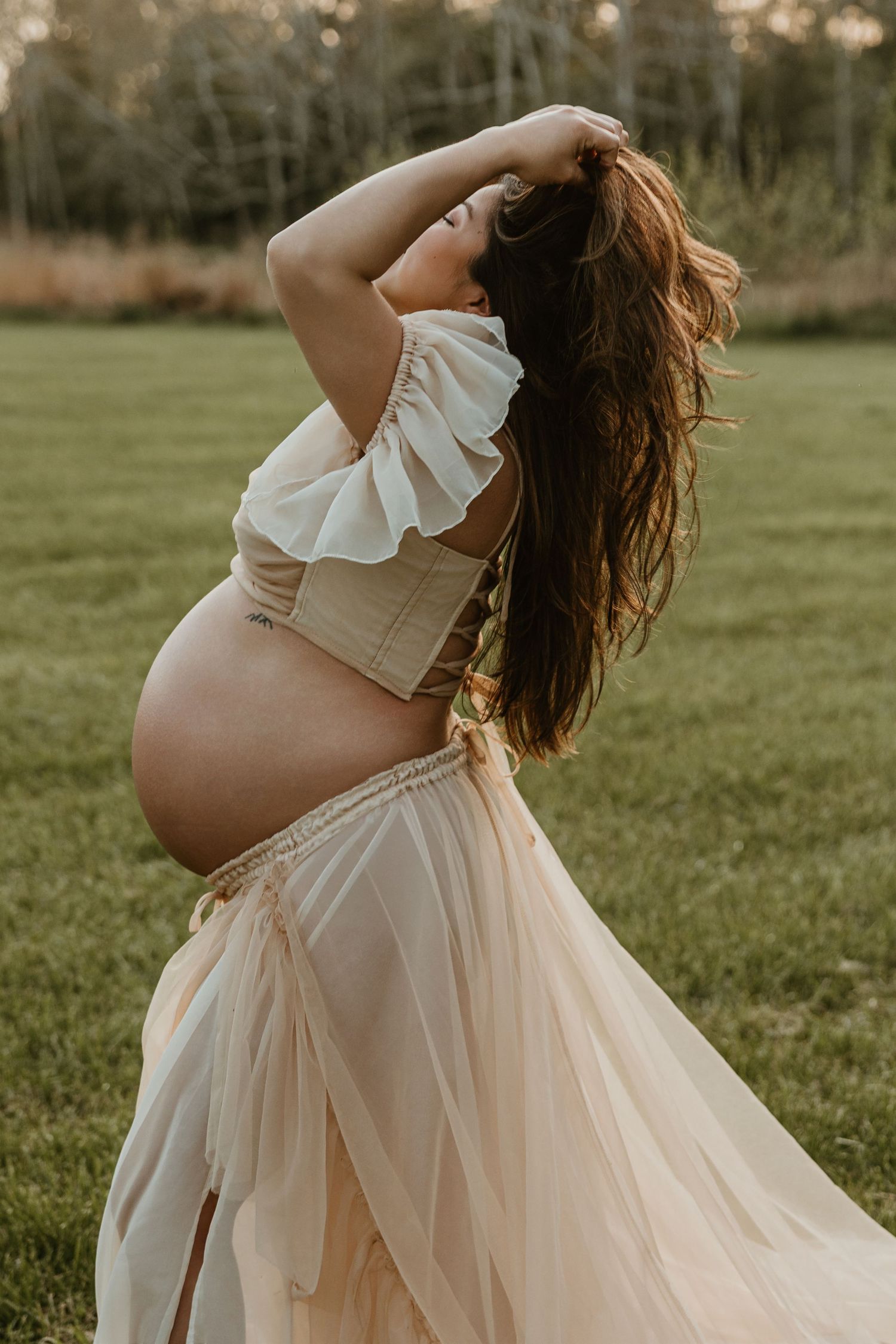 Pregnant woman in white ruffled crop top and sheer beige skirt posing outdoors in a green field at golden hour.