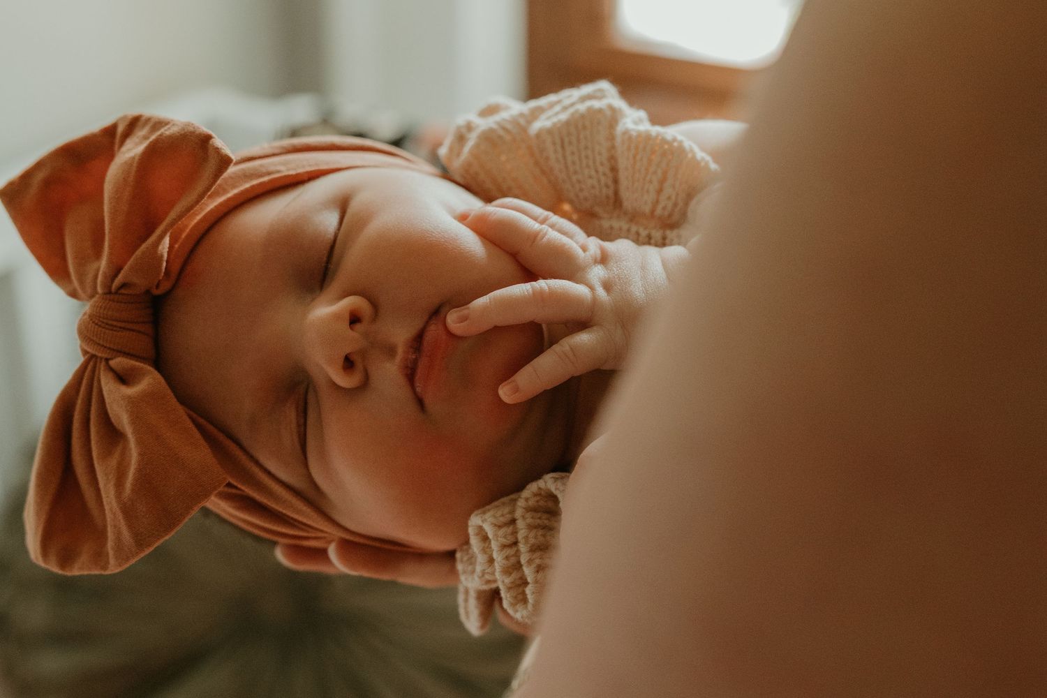 A sleeping newborn baby wearing an orange bow headband being held and breastfed by their mother.
