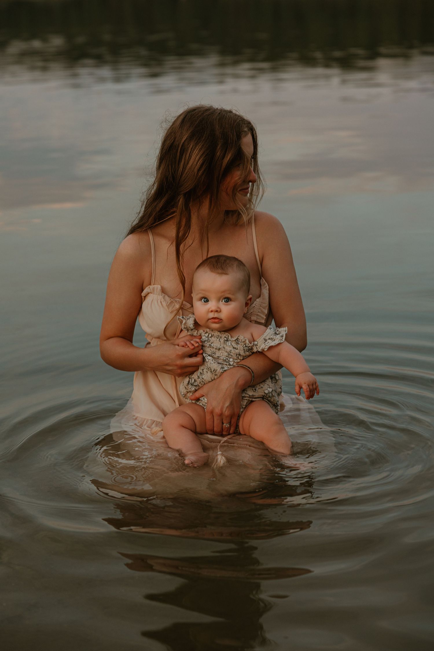 Mother holding baby while standing in calm lake water at sunset, both wearing light clothing in serene nature setting.