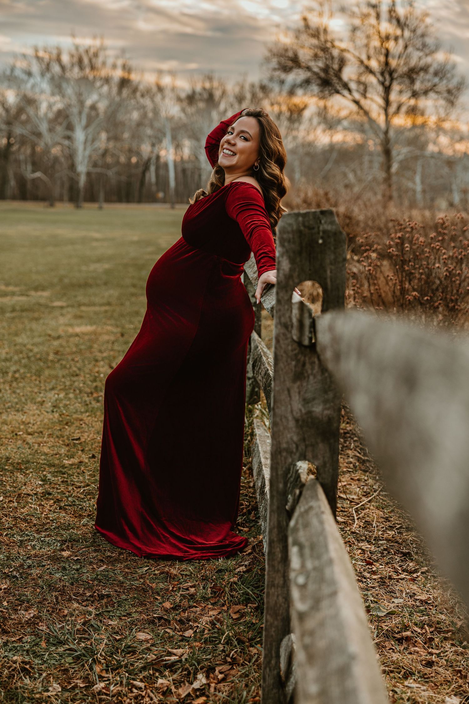 Person in a deep red dress poses expressively near weathered fence posts in an autumn field.