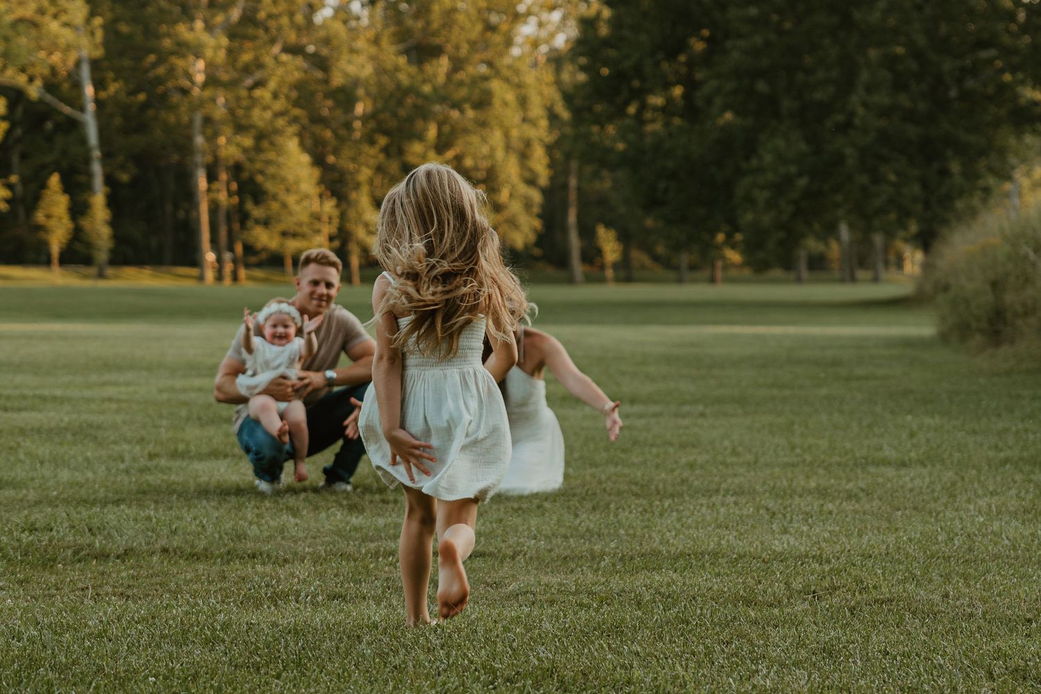 A young girl in a white dress runs barefoot across a lush green lawn toward her father and baby sibling.