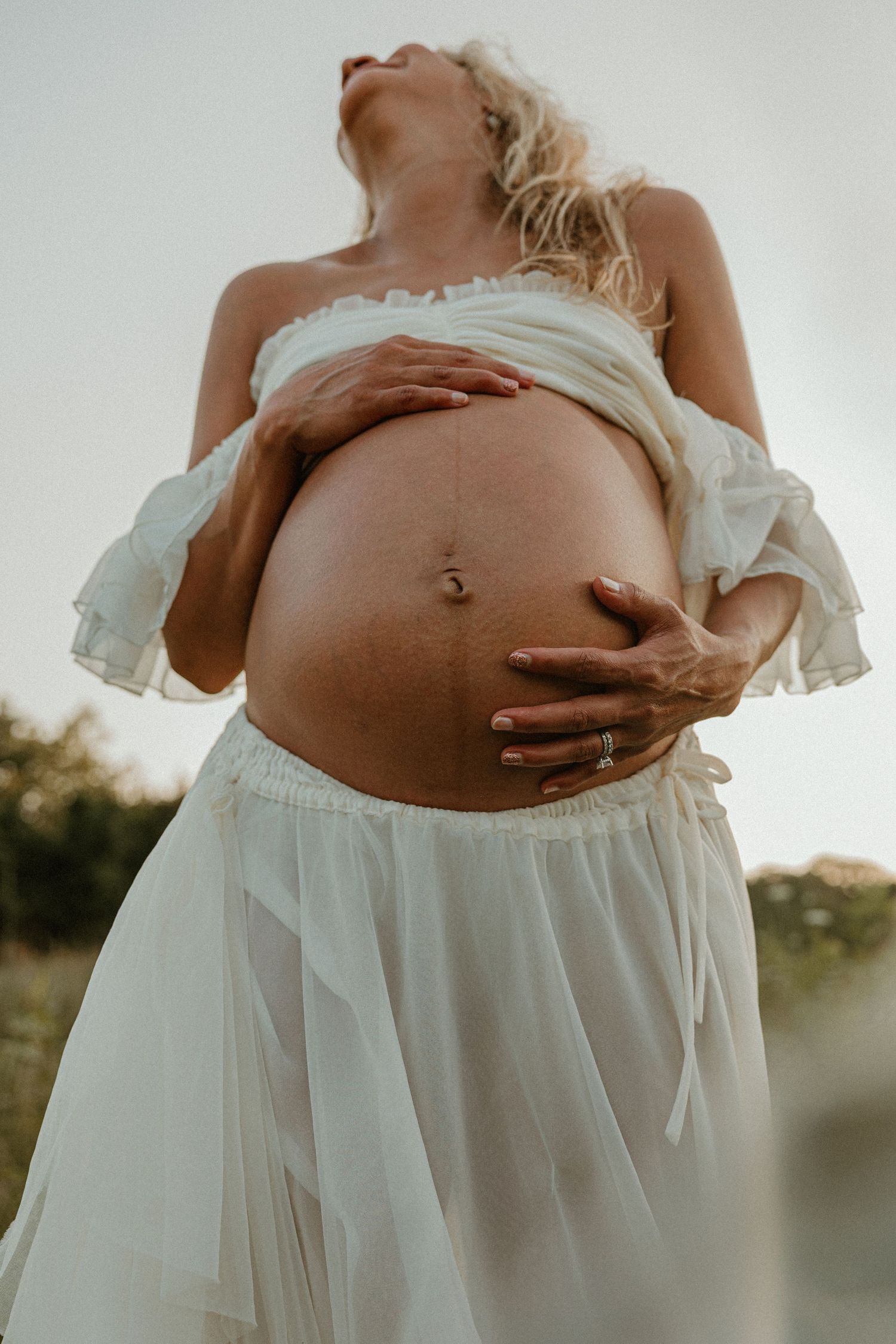 Pregnant woman in white dress cradling bare baby bump outdoors, maternity photography session.