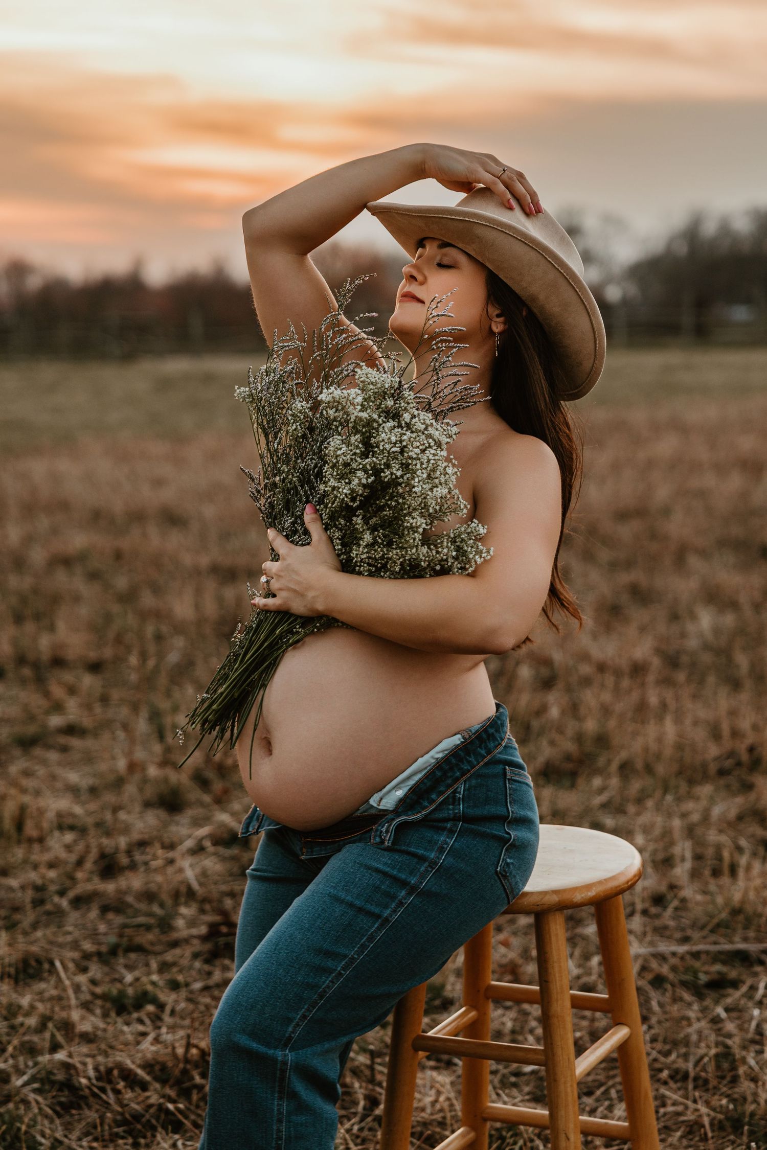 Pregnant woman in cowboy hat and jeans holds wildflowers at sunset in an open field during a maternity photo shoot.