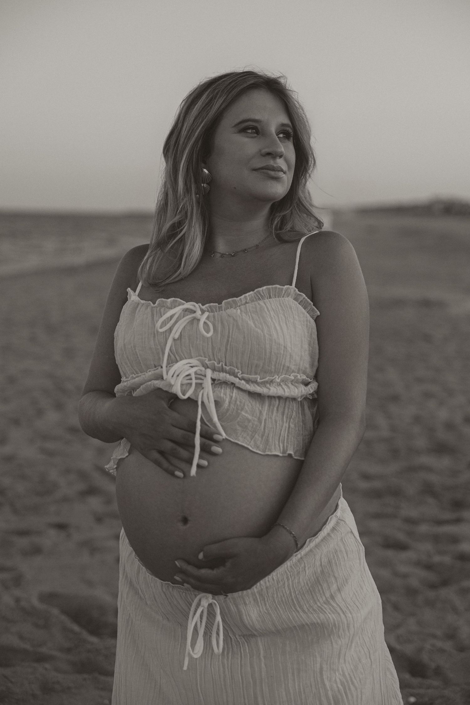 Black and white photo of a pregnant woman in a white crop top and skirt cradling her baby bump on a sandy beach.