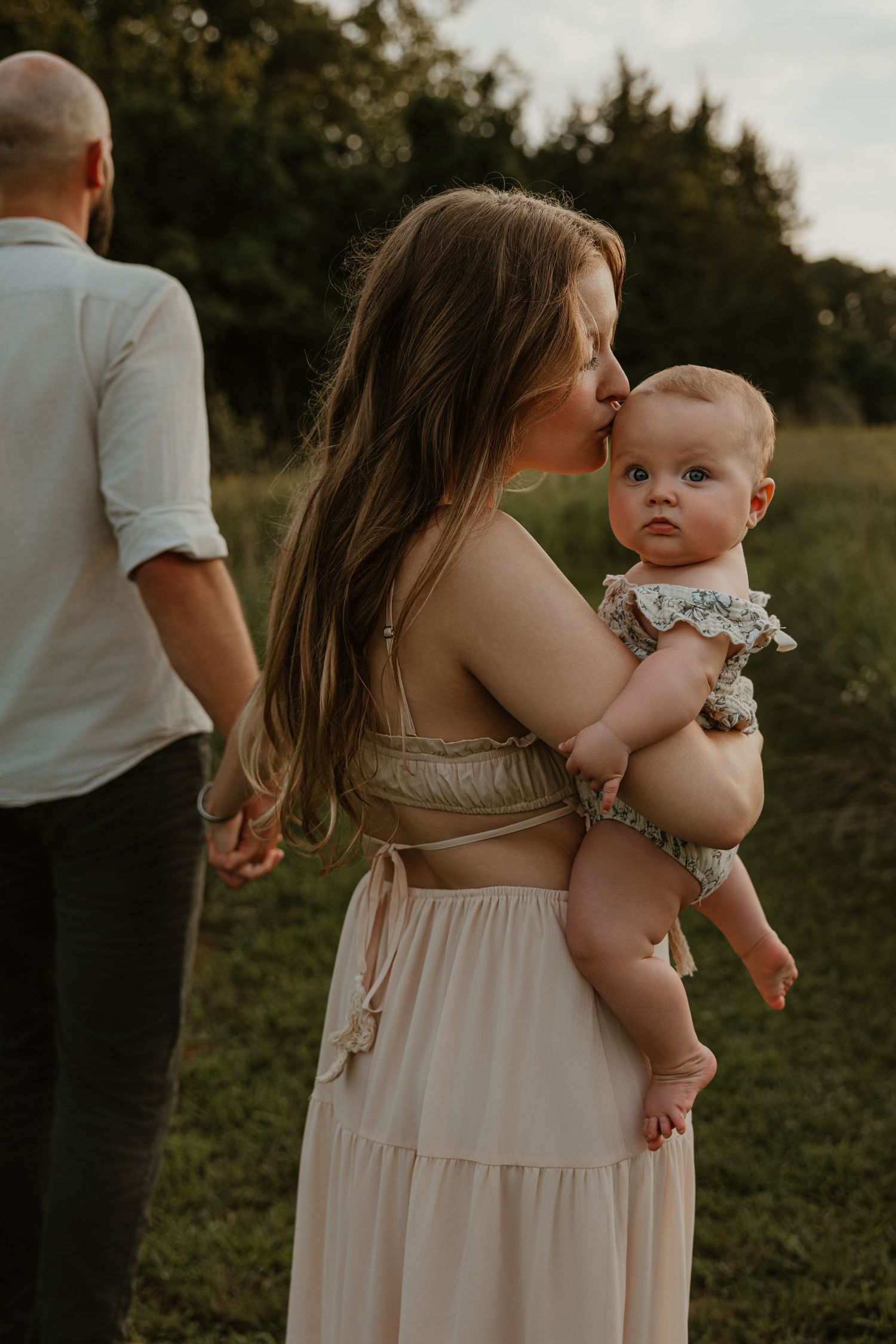 A mother kisses her baby while holding her in a sunlit field, as a man stands nearby holding her hand.