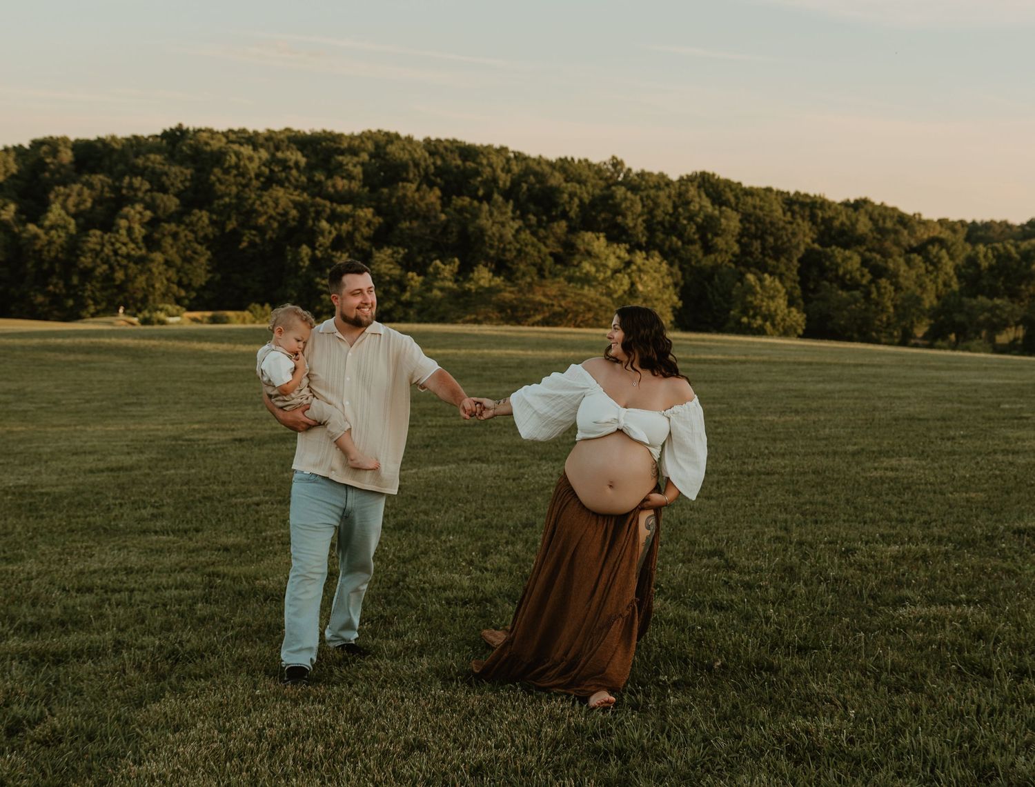 Pregnant woman in brown skirt holds hands with partner carrying toddler in a sunlit green field at golden hour.