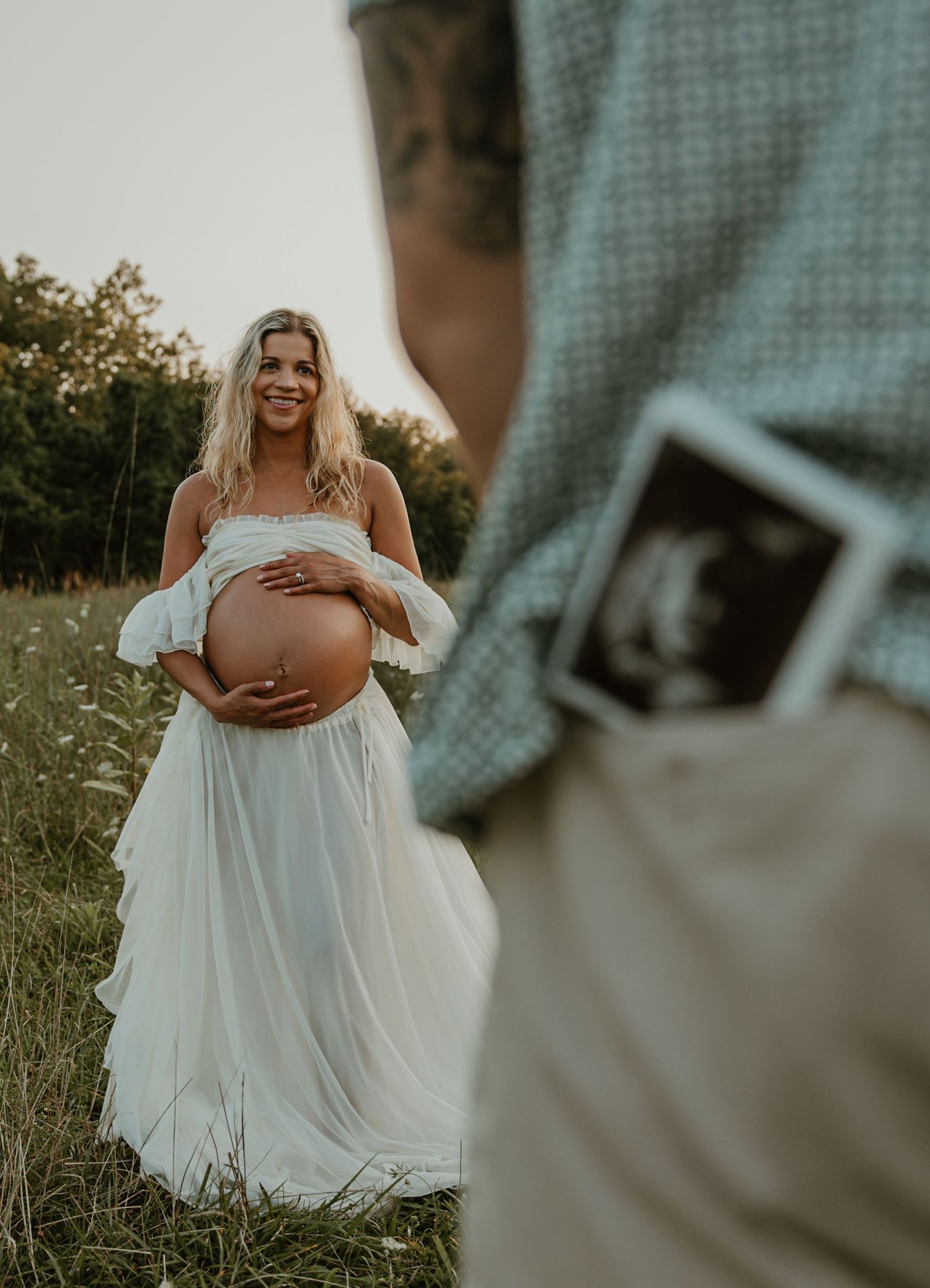 Pregnant woman in white dress holds baby bump in field while man holds ultrasound photo in foreground.
