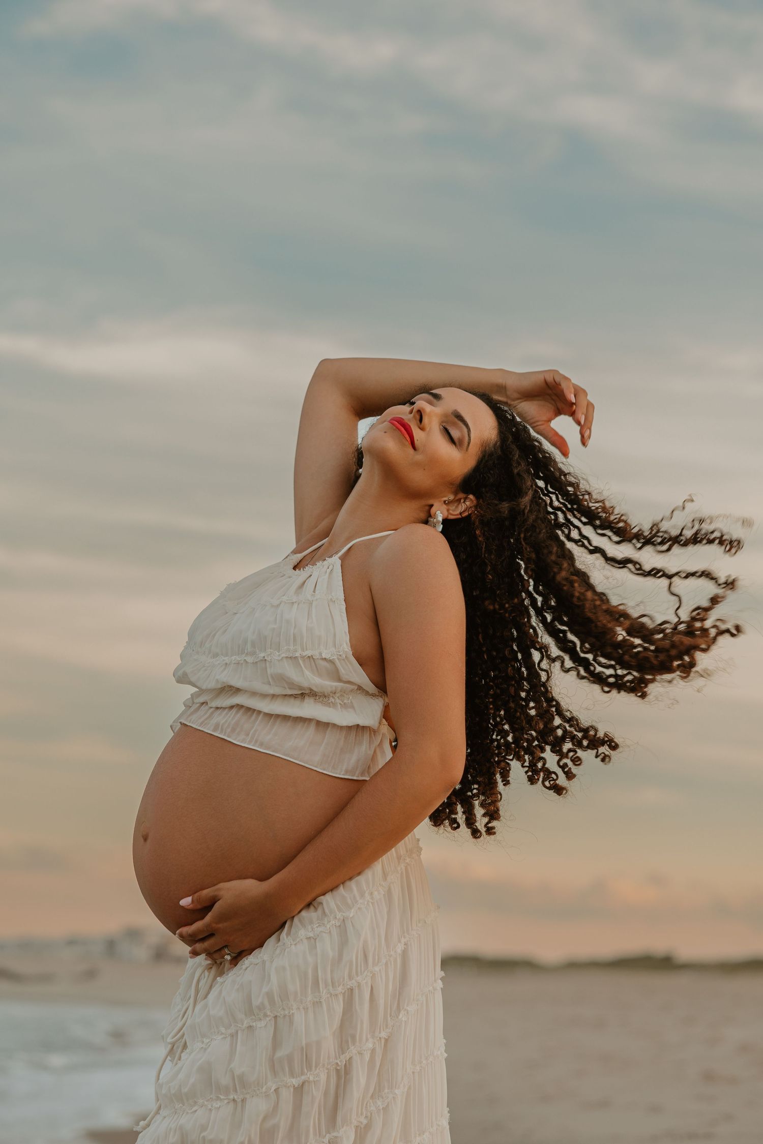 Pregnant woman in white dress poses gracefully on beach at sunset, cradling bump with curly hair blowing in wind.