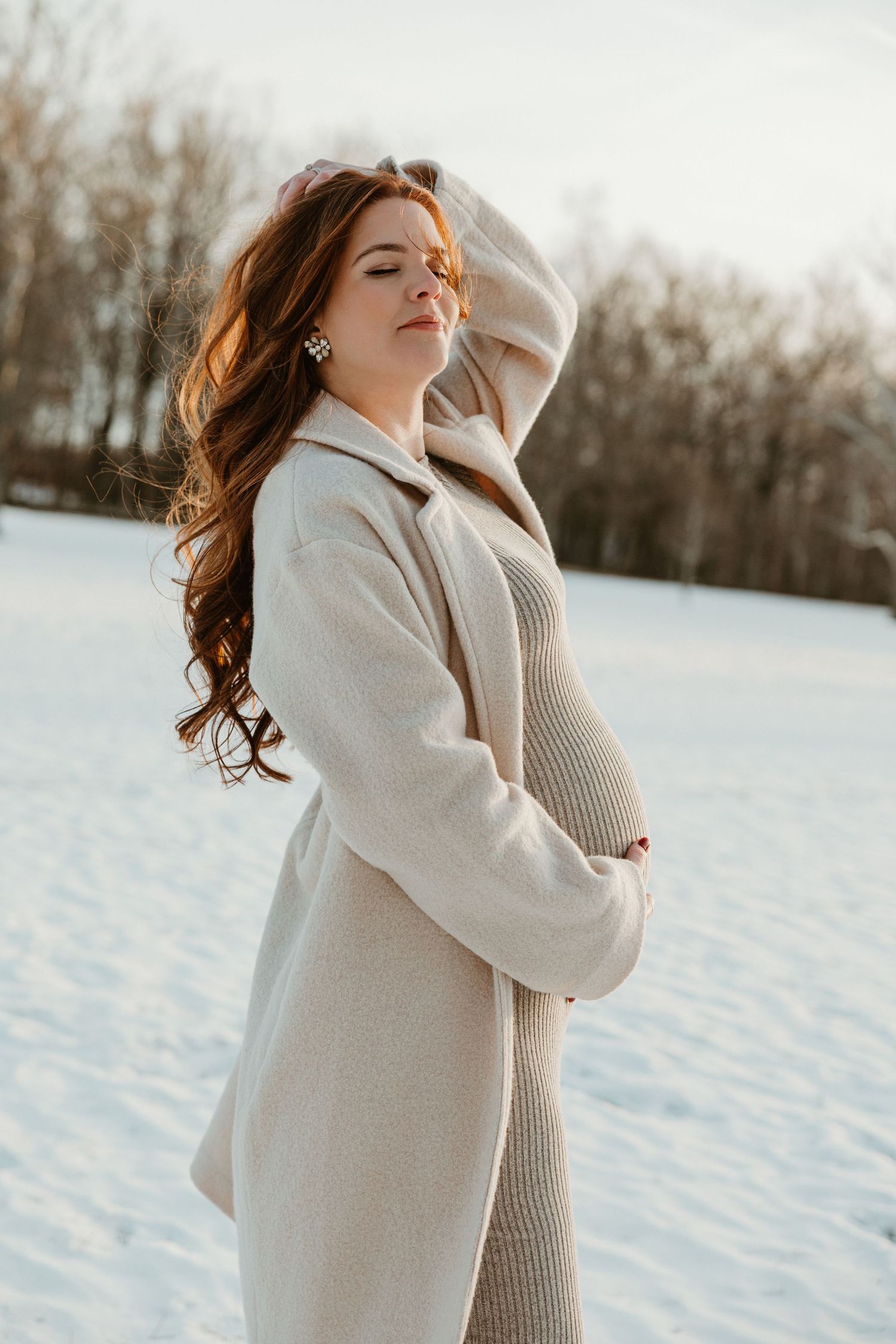 Expectant mother in cream sweater poses against snowy winter landscape.