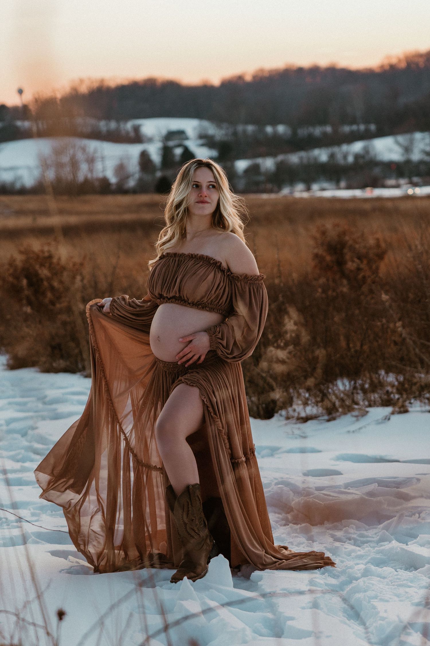 Ethereal winter maternity photos with a brown dress billowing in the snow-covered landscape.
